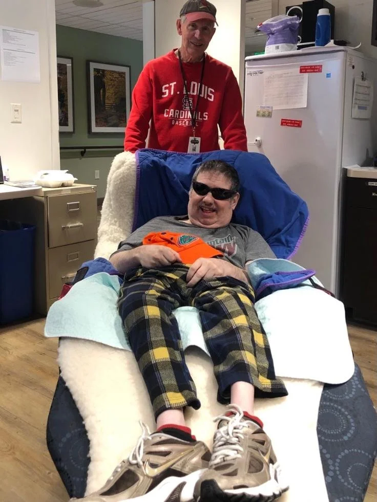 A young person in sunglasses and plaid pajama pants is sitting in a hospital bed, smiling and holding an orange piece of clothing. An older man in a red St. Louis Cardinals sweatshirt stands behind them, smiling and looking at the camera.