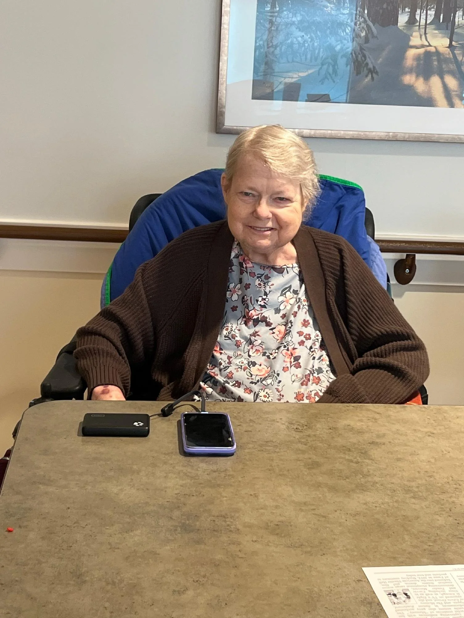 An elderly woman with short gray hair sitting at a table in a room, wearing a floral top and a brown cardigan, smiling at the camera. There are two mobile phones on the table in front of her. The background has a framed picture of a forest scene and 