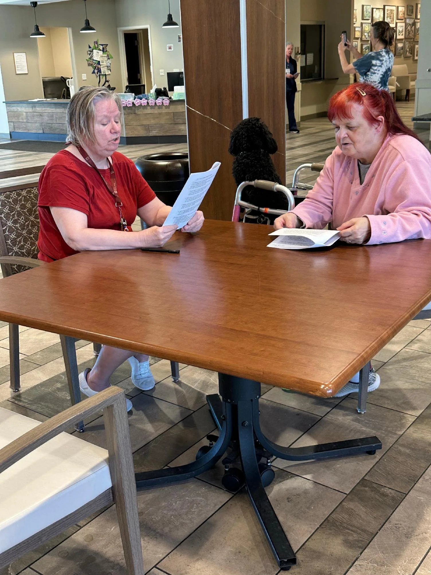 Two women with light skin sitting at a wooden table in a lobby or waiting area, looking at papers. One is wearing a red shirt and the other a pink sweater. A black poodle dog is sitting on a chair between them. In the background, a person with a pony