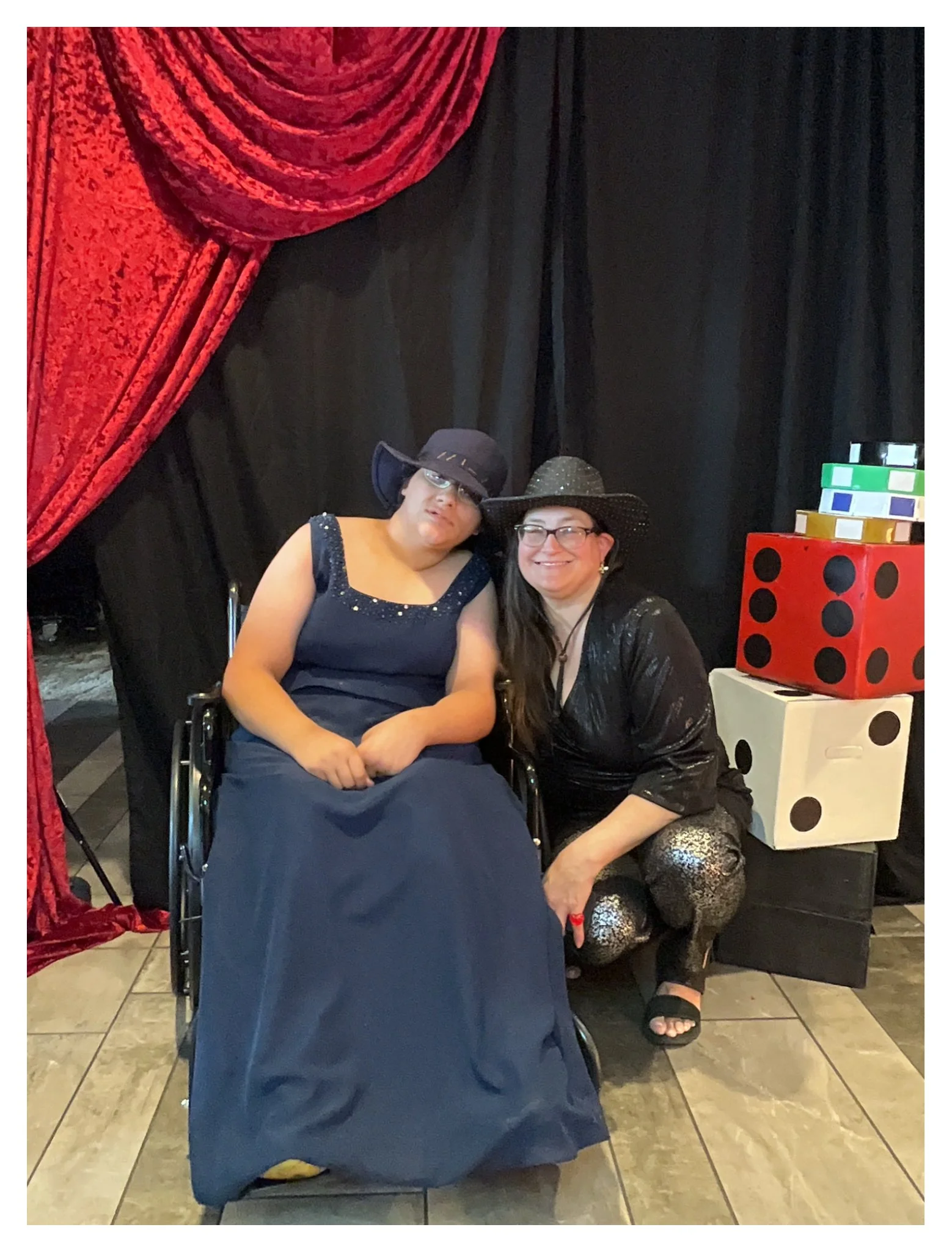 Two women posing together at a party, wearing hats and formal dresses, with large decorative dice in the background.