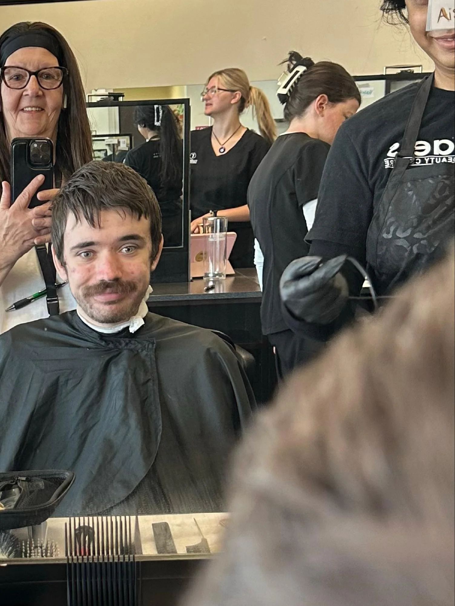 People in a salon, with a woman holding a phone, a man with a barber cape, and two women working behind the counter.