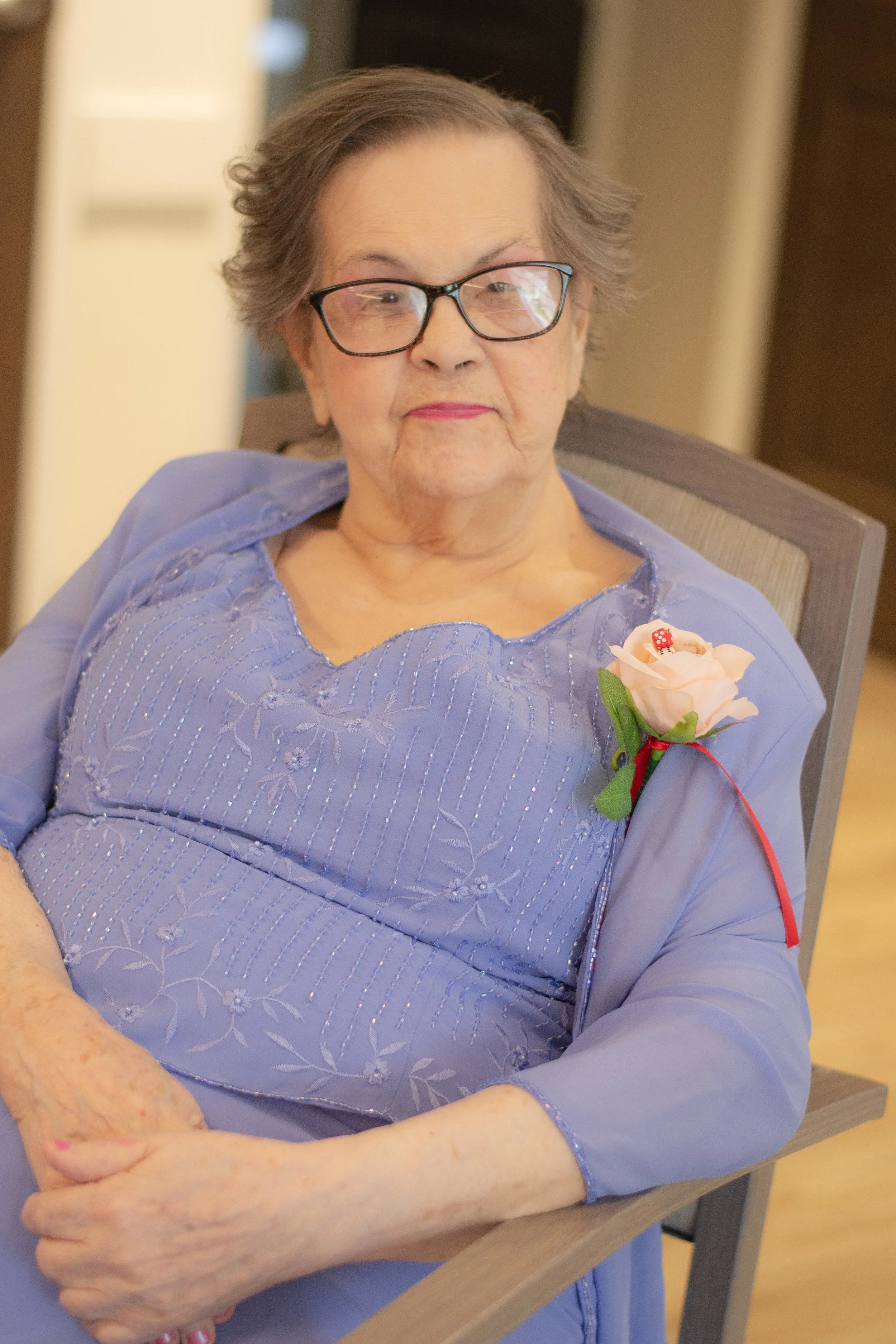 An elderly woman with glasses, wearing a lavender dress with embroidery, is sitting at a table with her arms crossed. She has a flower with a ladybug on it pinned to her dress and appears to be smiling softly.