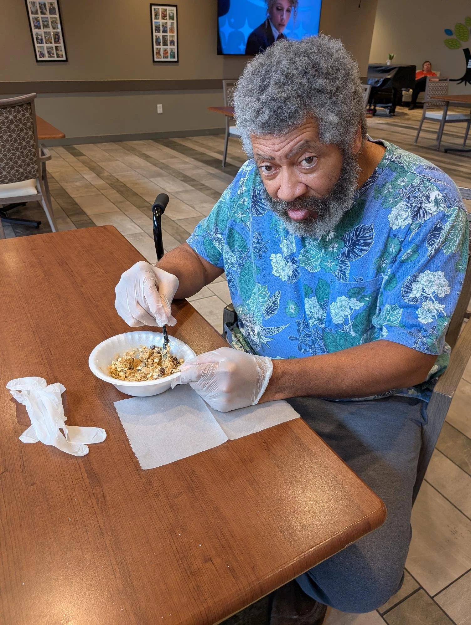 An elderly man with gray hair and a beard, wearing a blue floral shirt, is sitting at a wooden table in a restaurant. He is holding a spoon and eating from a bowl of cereal. He is wearing white gloves. In the background, there is a television on the 