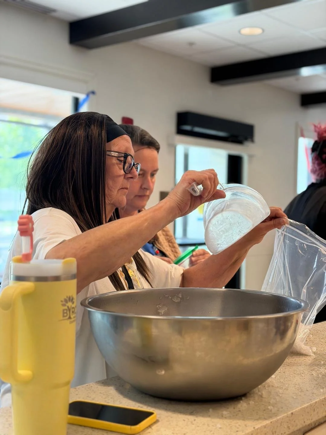 Women working with a large metal mixing bowl, pouring a clear substance from a plastic container, in an indoor setting with natural light and a window in the background.
