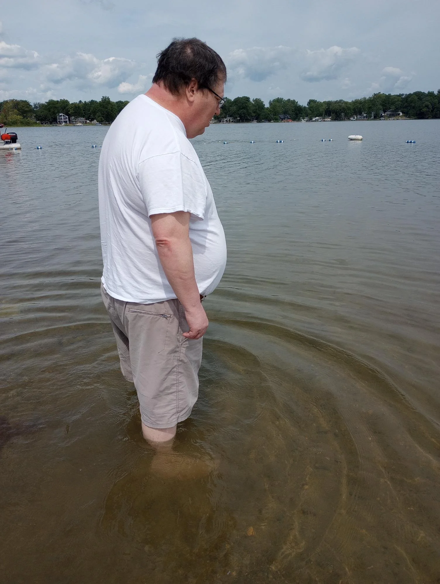 A middle-aged man wearing a white T-shirt and khaki shorts stands in shallow water at a lakeside, looking down at the water.
