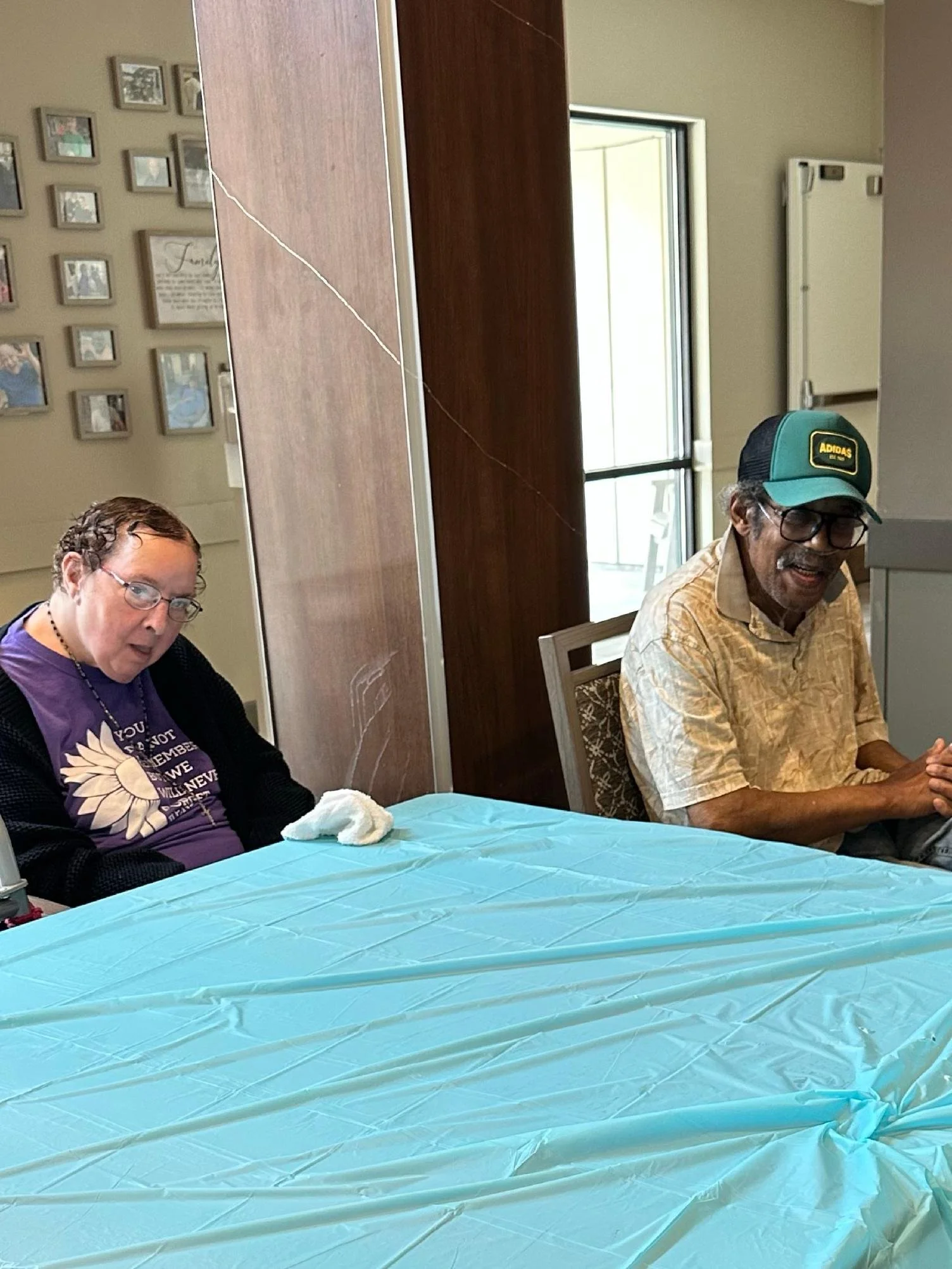 Two people sitting at a table covered with a light blue tablecloth in a room with framed photos on the wall. The woman on the left has glasses and curly hair, wearing a dark top with a purple shirt underneath, and is looking down. The man on the righ