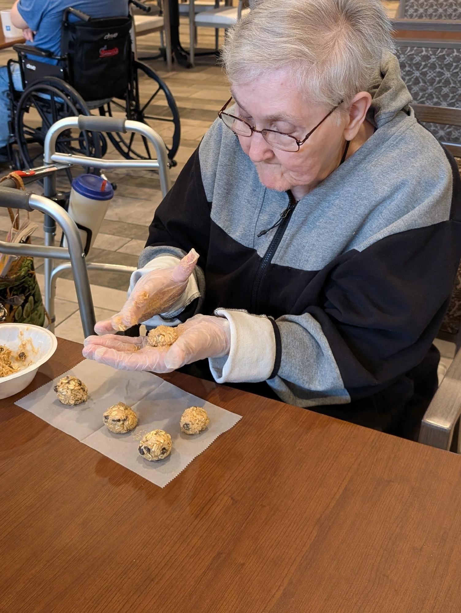 An elderly woman with glasses is wearing gloves and preparing to place cookie dough balls on parchment paper on a wooden table, with bowls of cookie ingredients nearby.