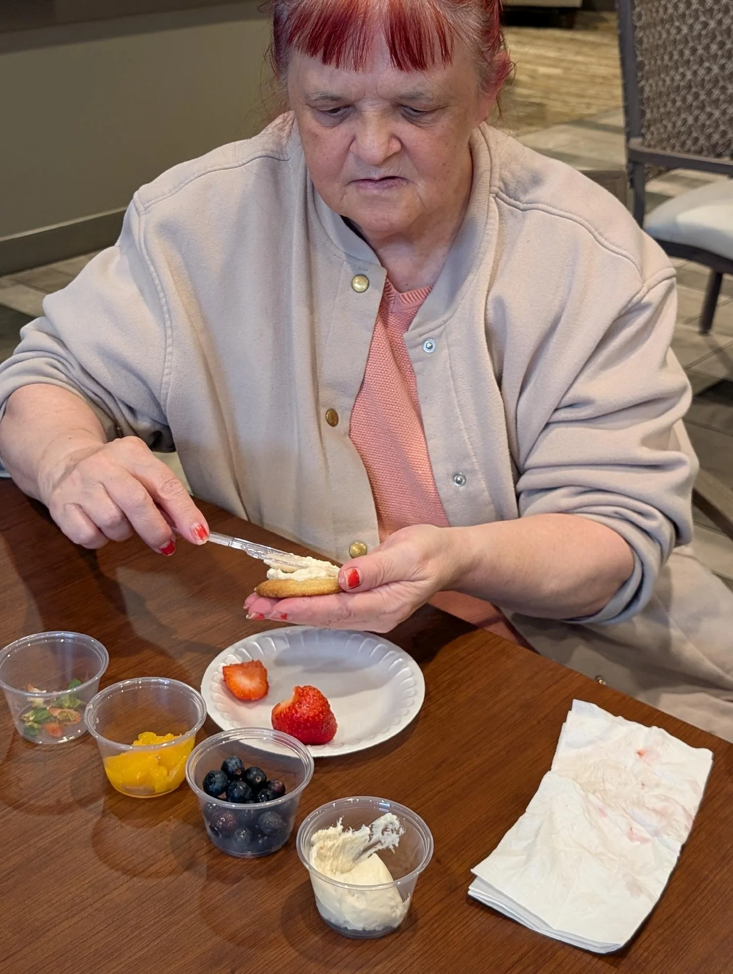 An elderly woman with red hair and painted nails playing on a cookie with cream and strawberries. There are strawberries, blueberries, and various toppings and creams on the table.