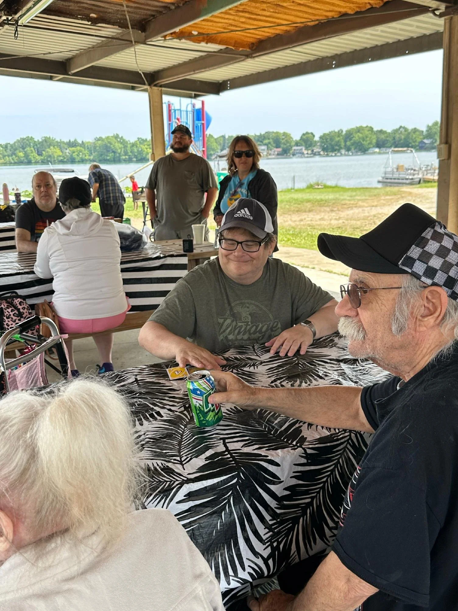People gathered under a pavilion near a lake, sitting at a table with a black and white tropical leaf pattern tablecloth. Some are standing, and some are sitting, with a view of water, a boat, and a playground in the background. One person is holding