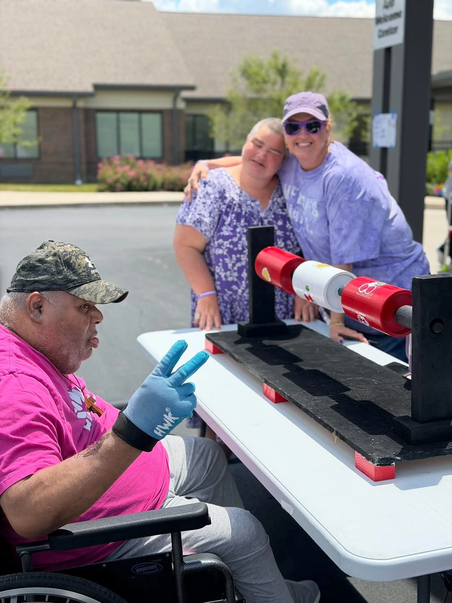 A man in a wheelchair wearing a pink shirt and gray pants, making a peace sign with his hand, sitting at an outdoor table with two women, one of whom has her shoulder on the other woman's shoulder. The women are smiling, and there's a black table wit