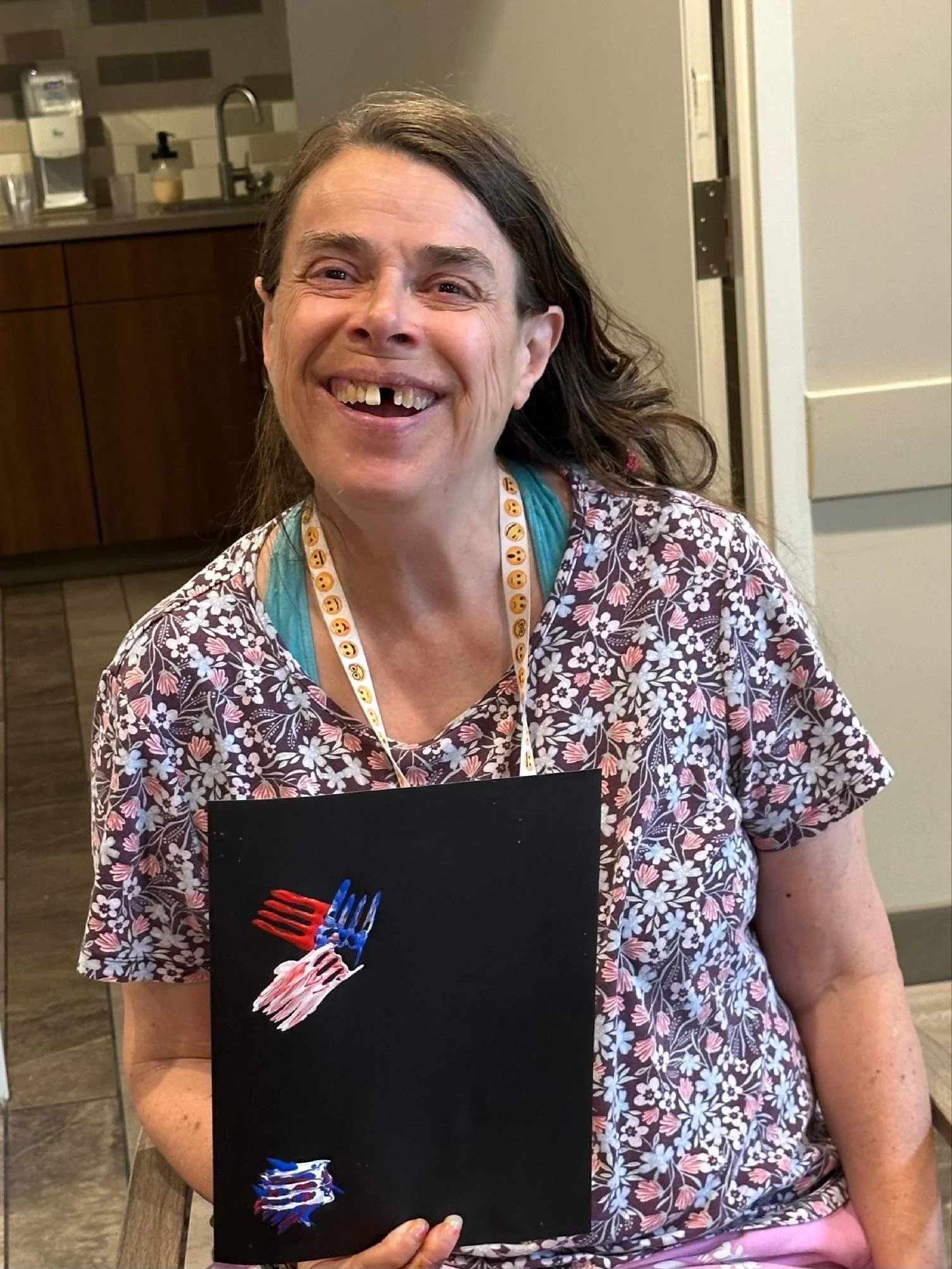 A smiling woman with missing front teeth holding a black poster with a colorful fork illustration, wearing a floral shirt and a badge with smiley face emojis around her neck, standing in a kitchen.