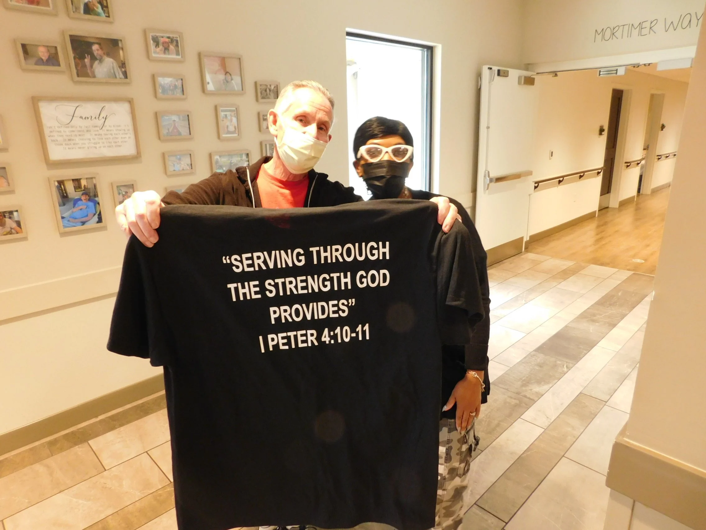 Two women wearing face masks standing indoors, one holding a black cloth banner with religious text from 1 Peter 4:10-11, in front of a wall with photo frames and a window.