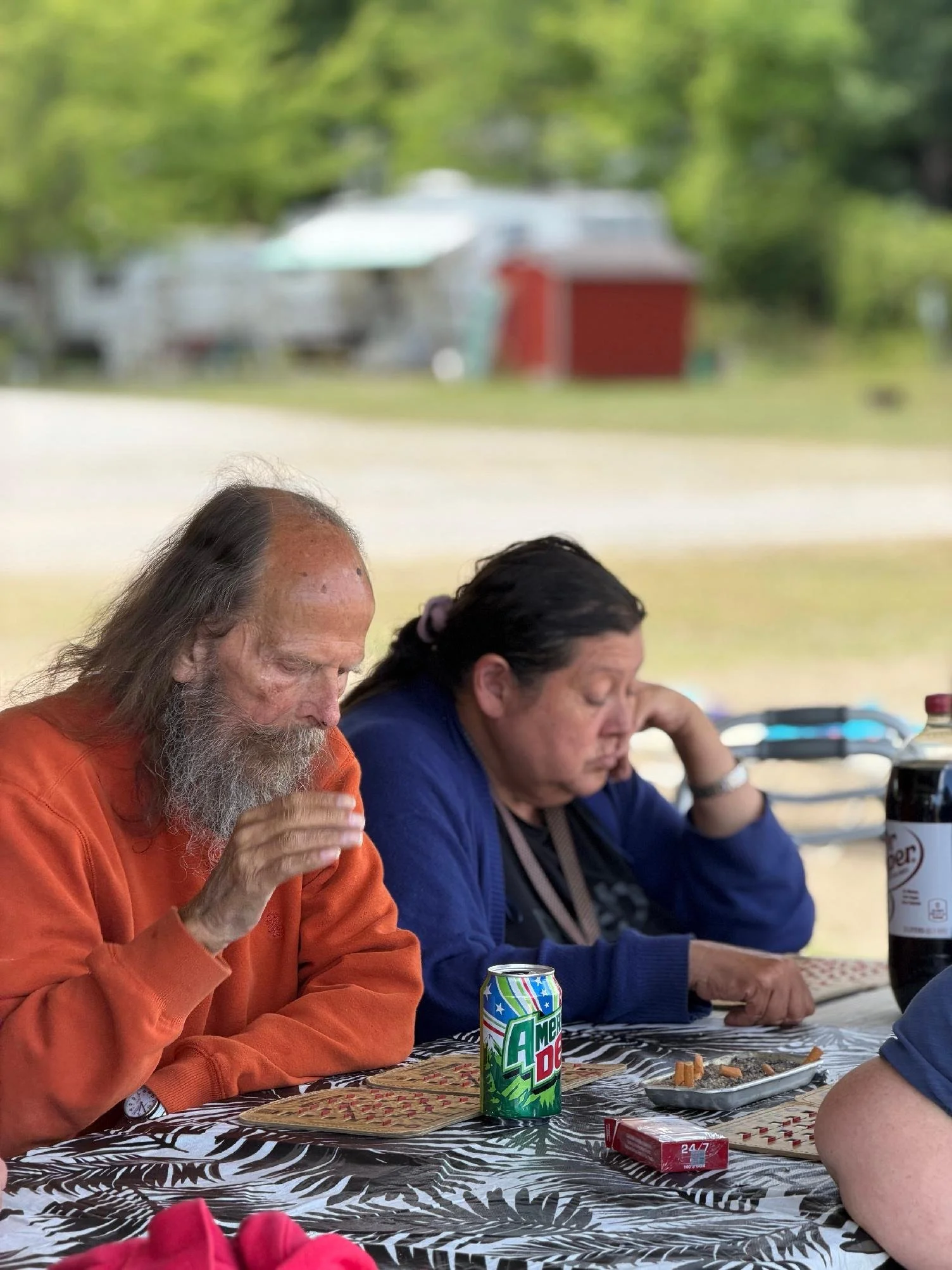 Two people are seated at a table outdoors during daytime. The person on the left has long gray hair, a beard, and is wearing an orange sweatshirt. The person on the right has dark hair tied back, wearing a dark blue top. There is a can of Mountain De
