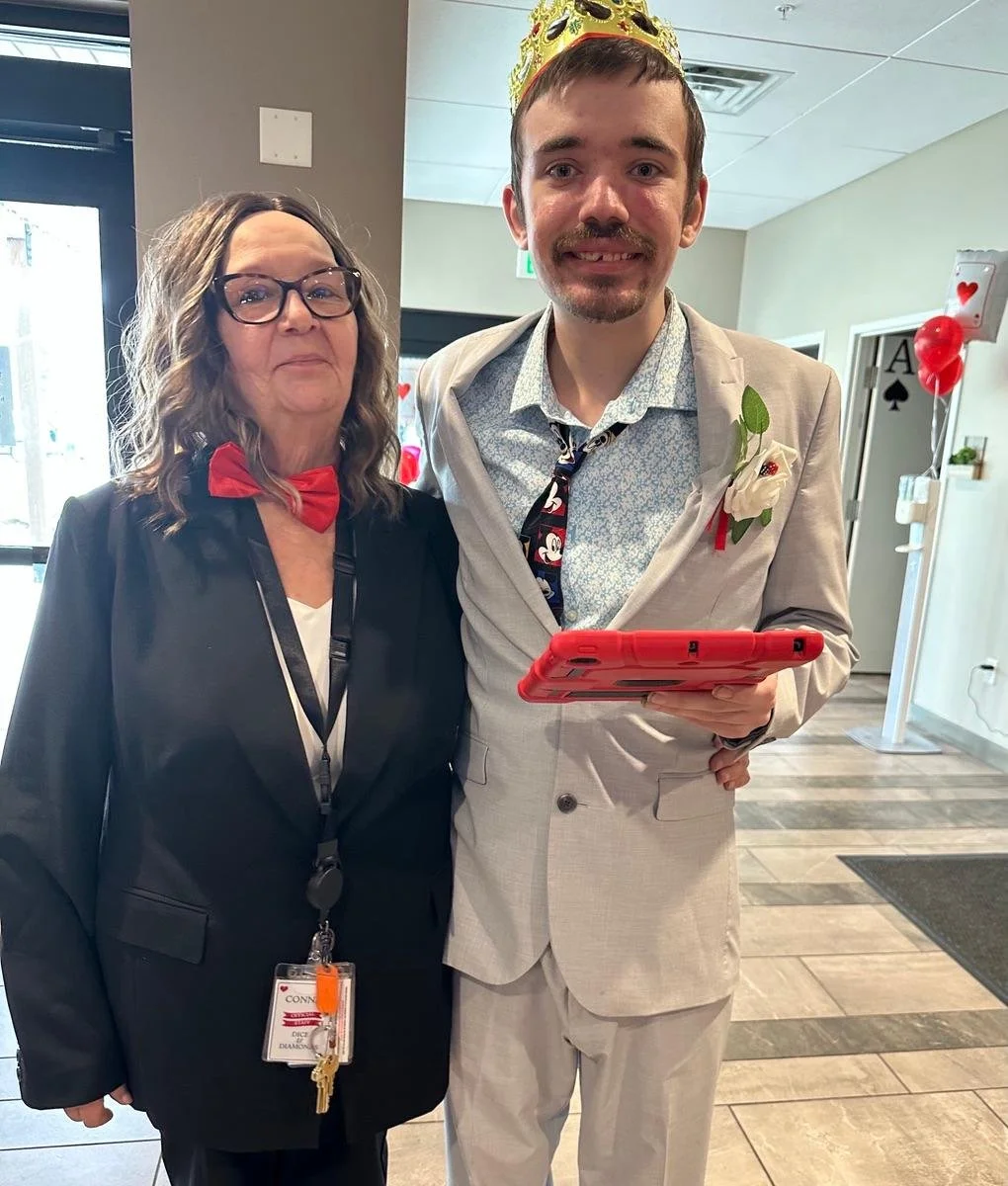 A man and woman stand together indoors during a celebration. The man wears a light gray suit, a white shirt, and a Mickey Mouse tie, with a white rose boutonniere. He has a gold crown on his head and holds a tablet. The woman wears black glasses, a b