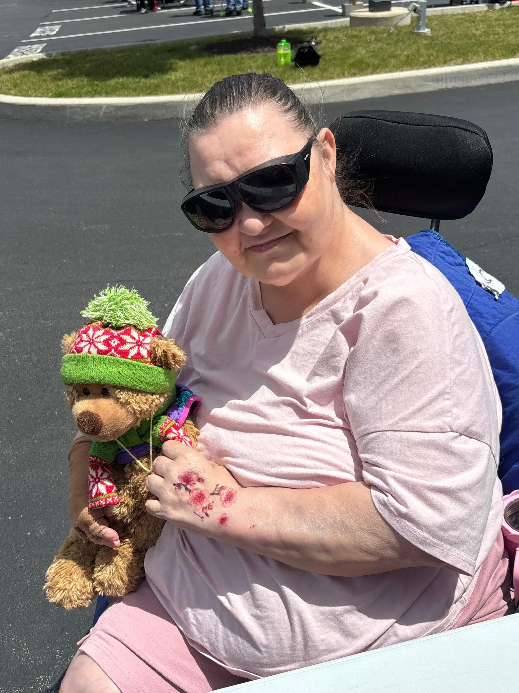 A woman with dark hair tied back, wearing black sunglasses and a light pink shirt, is sitting on a motorized wheelchair outdoors on a road with a grassy median behind her. She is smiling and holding a small decorated teddy bear wearing a Christmas sw