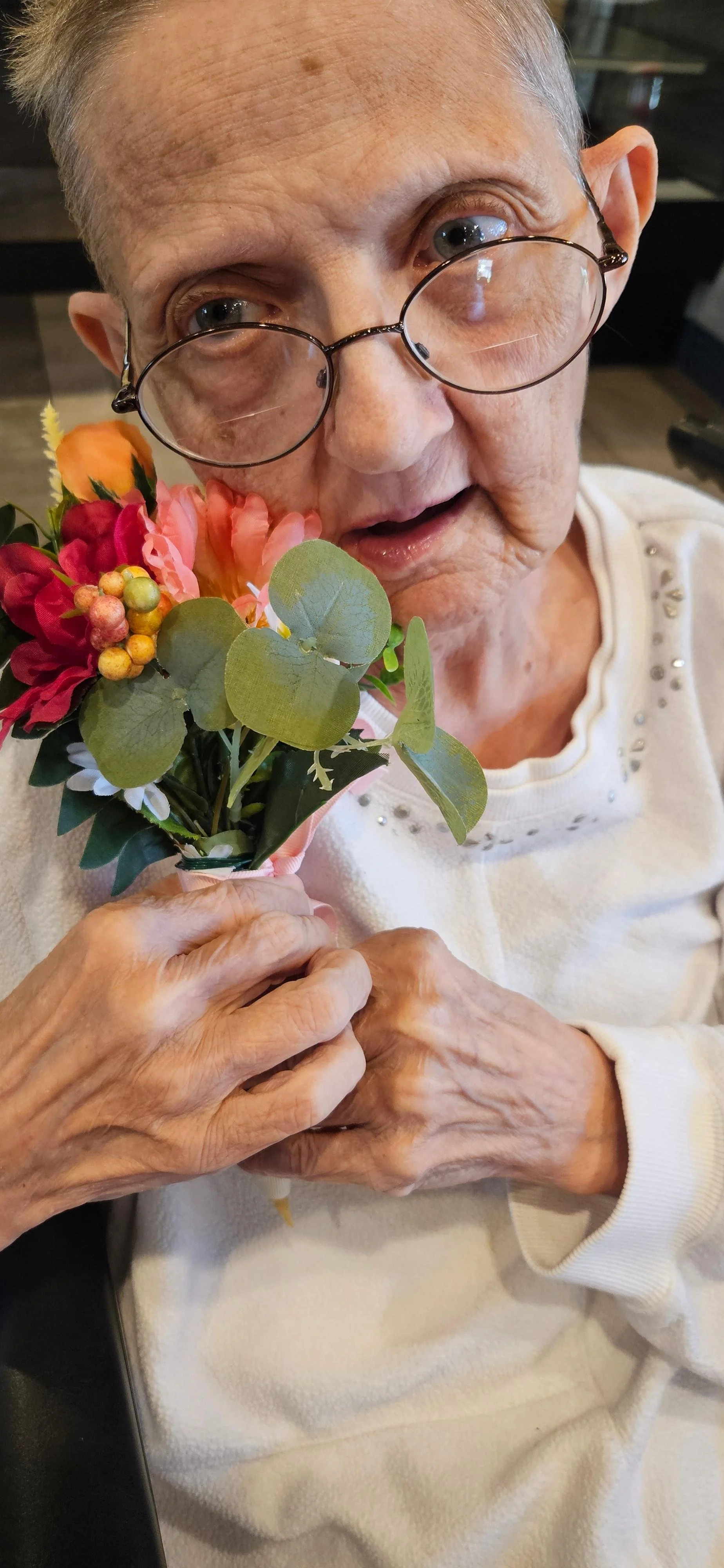 An elderly woman with glasses holding a small bouquet of colorful flowers and greenery, wearing a cream-colored sweater with decorative studs near the collar.