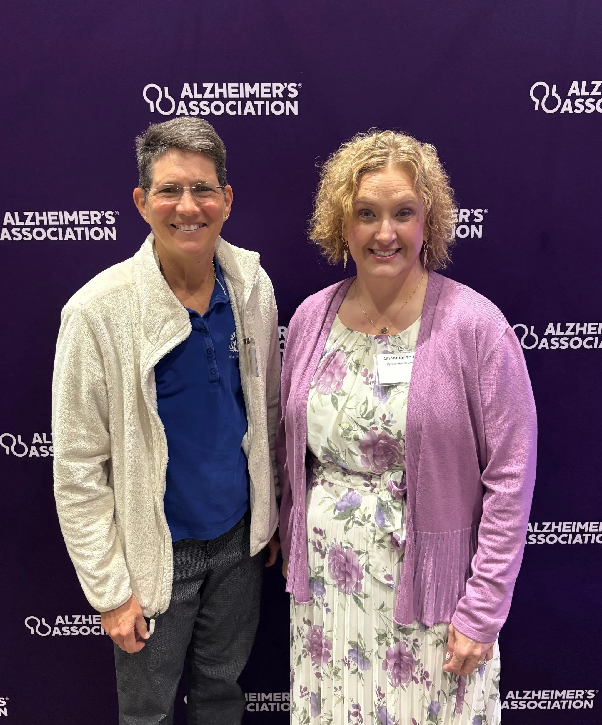 Two women standing side by side in front of a purple background with the Alzheimer’s Association logo. The woman on the left has short gray hair, glasses, and is wearing a white fleece jacket over a blue shirt. The woman on the right has curly blond 