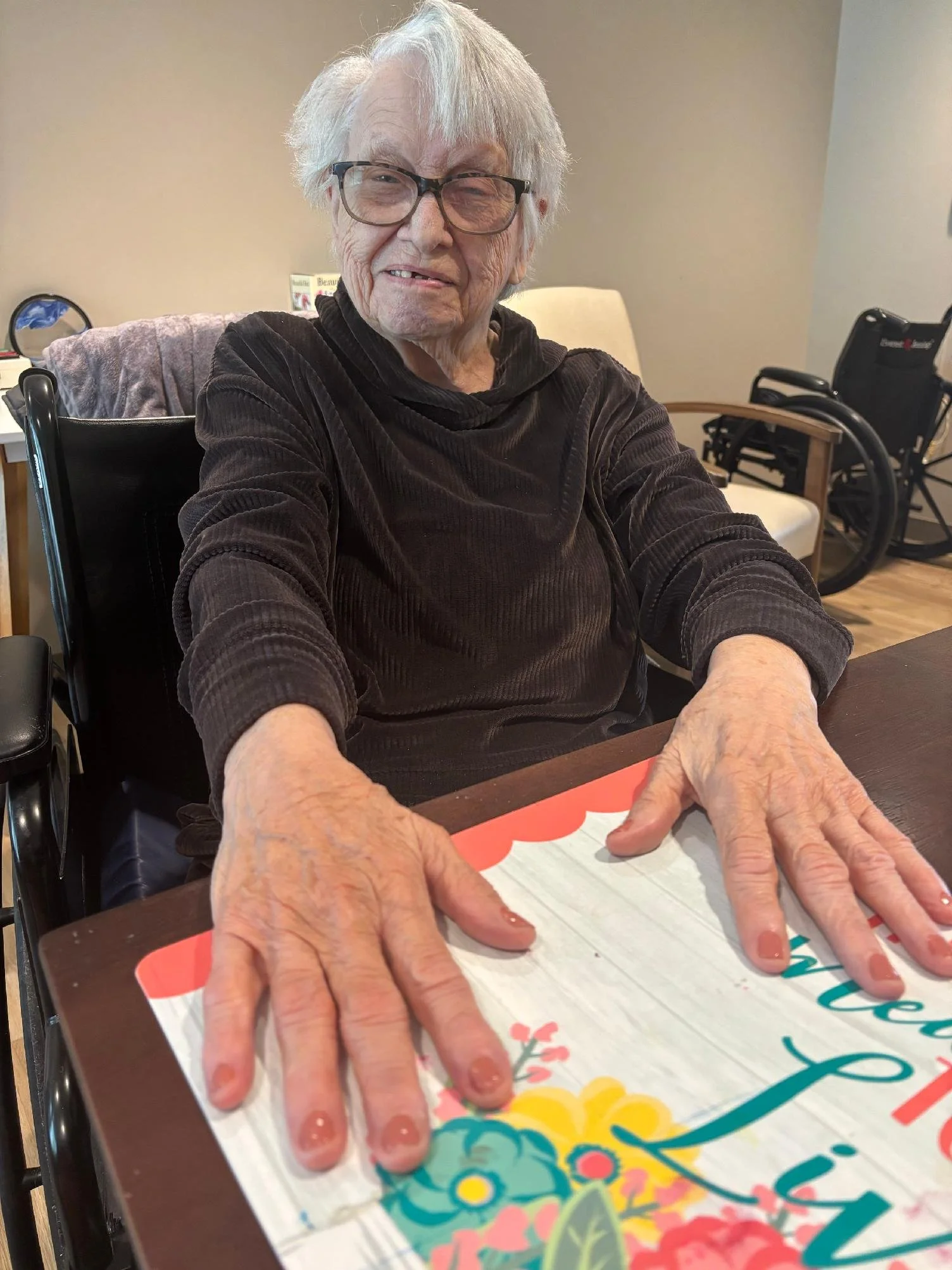 An elderly woman with white hair, glasses, and a dark velvety top sitting at a table showing off her freshly painted purple. She is in a wheelchair, and another wheelchair can be seen in the background, along with a beige wall and a small side table.