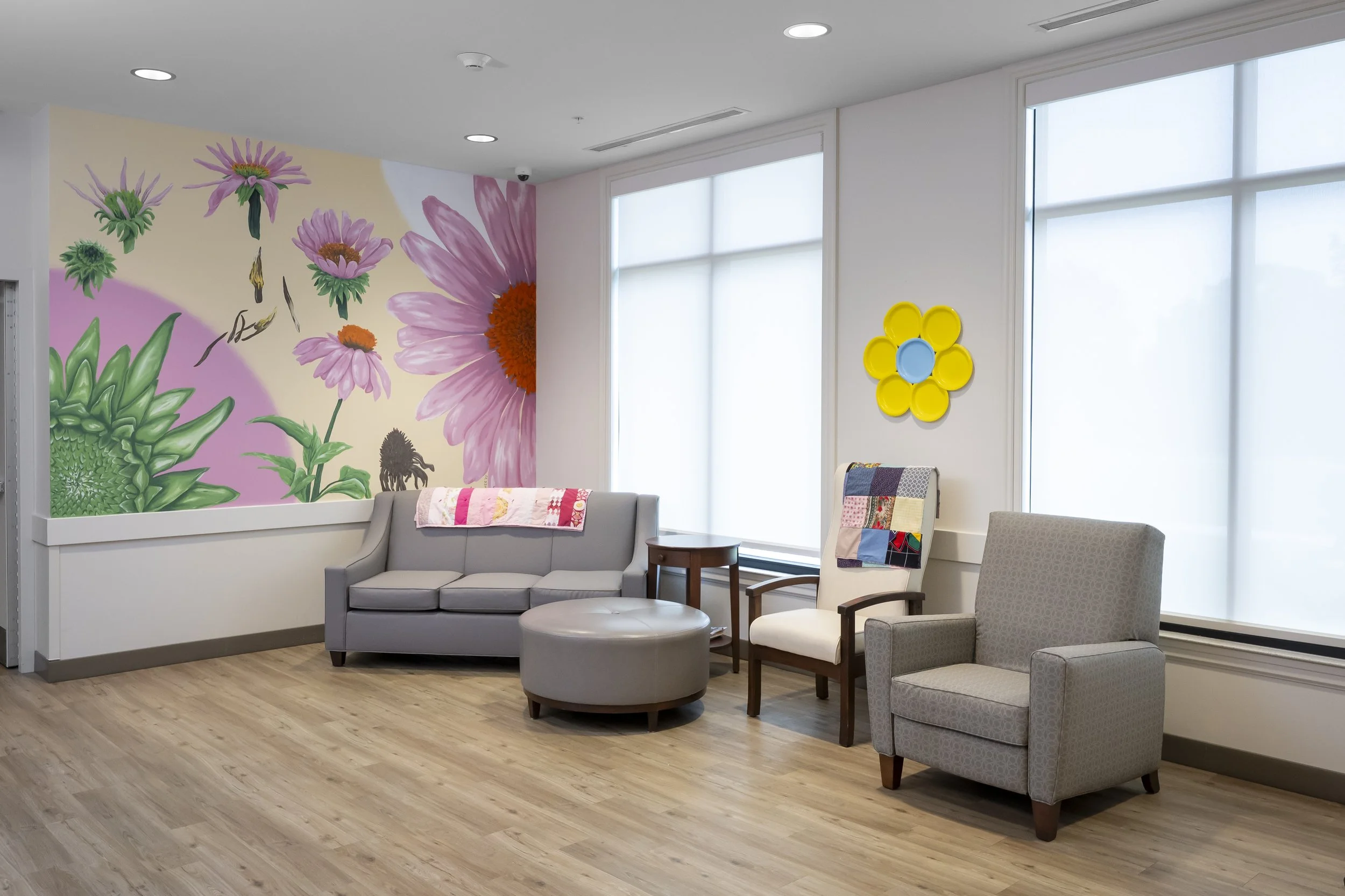 A waiting room with light wood flooring, a floral mural on one wall, a gray sofa with a pink quilt, two armchairs, and a large window with blinds.
