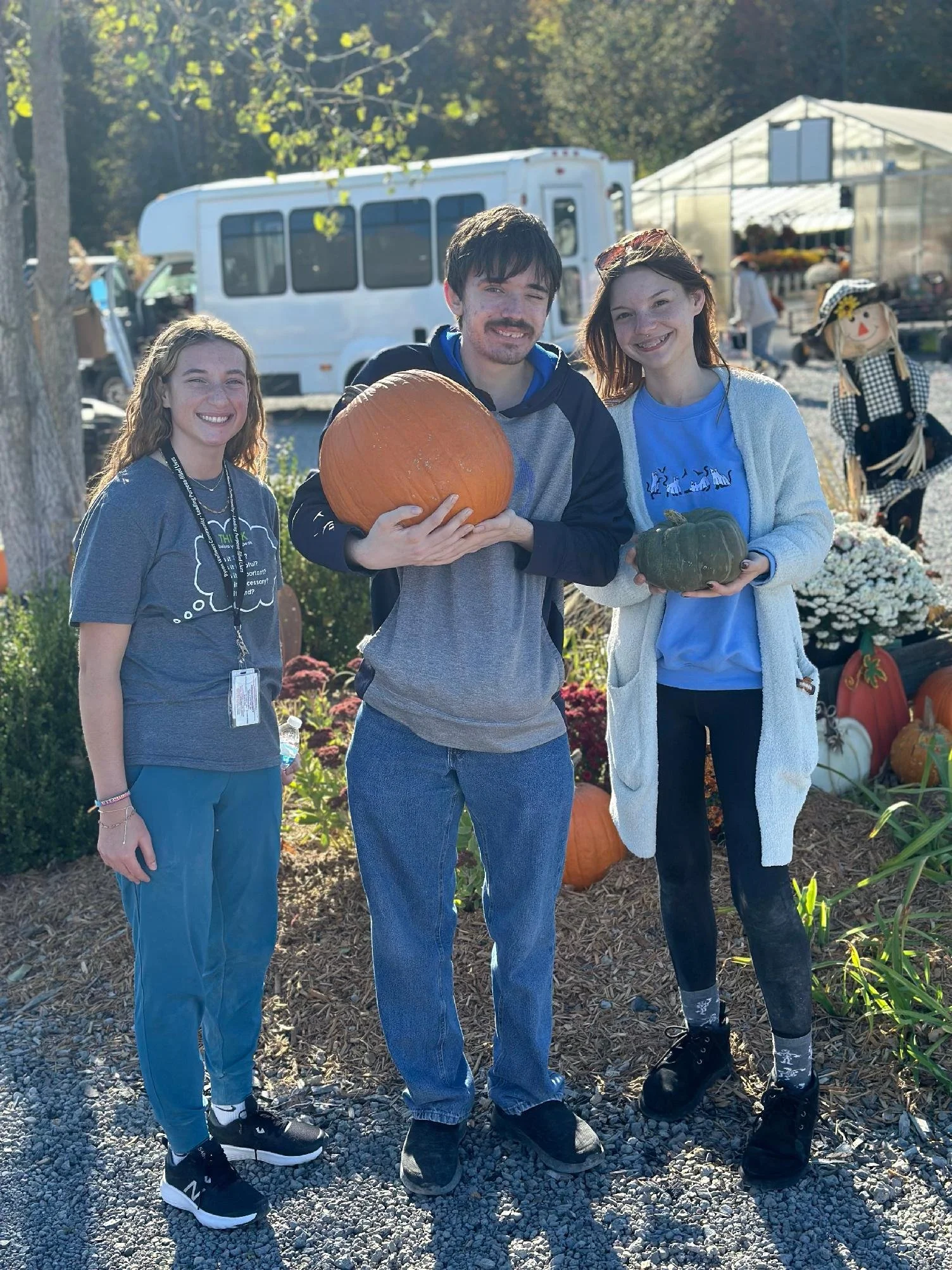 Three people standing outdoors, holding pumpkins, at a pumpkin patch or farm, with a white bus and market stall in the background.