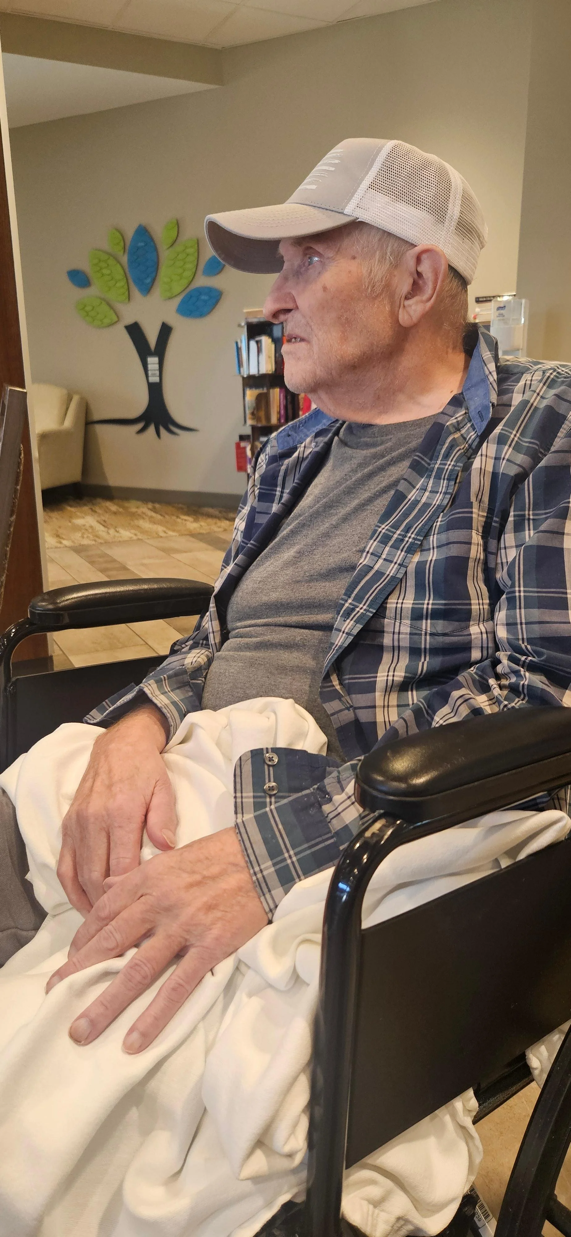 An elderly man sitting in a wheelchair, wearing a gray t-shirt, plaid shirt, white pants, and a baseball cap, in a room with a bookshelf and a wall decoration resembling a tree.