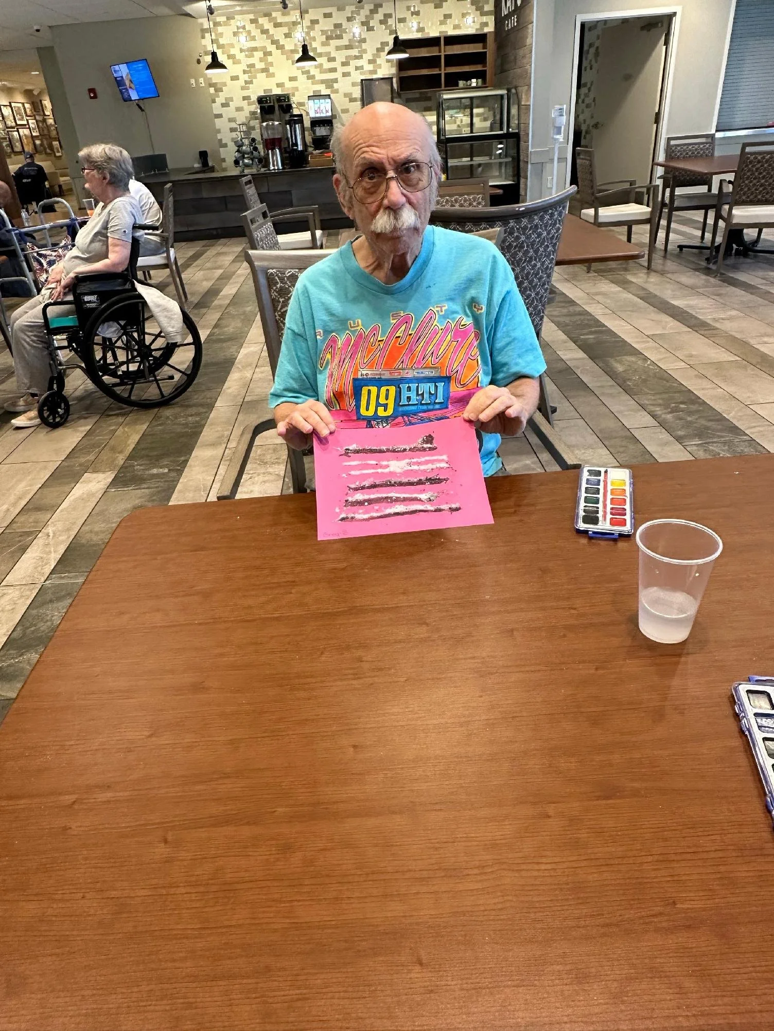 An elderly man with glasses, a mustache, and a bald head, sitting at a wooden table in a cafe. He is wearing a bright blue T-shirt with colorful text. In front of him is a pink paper with some lines on it, and a watercolor palette. To the left in the
