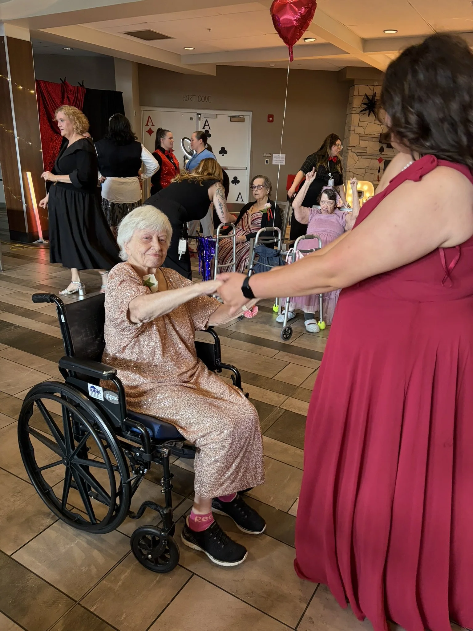 Group of women at a social gathering, some standing and some seated in wheelchairs, with decorations including red heart-shaped balloons, playing cards on the wall, and illuminated letter signs.