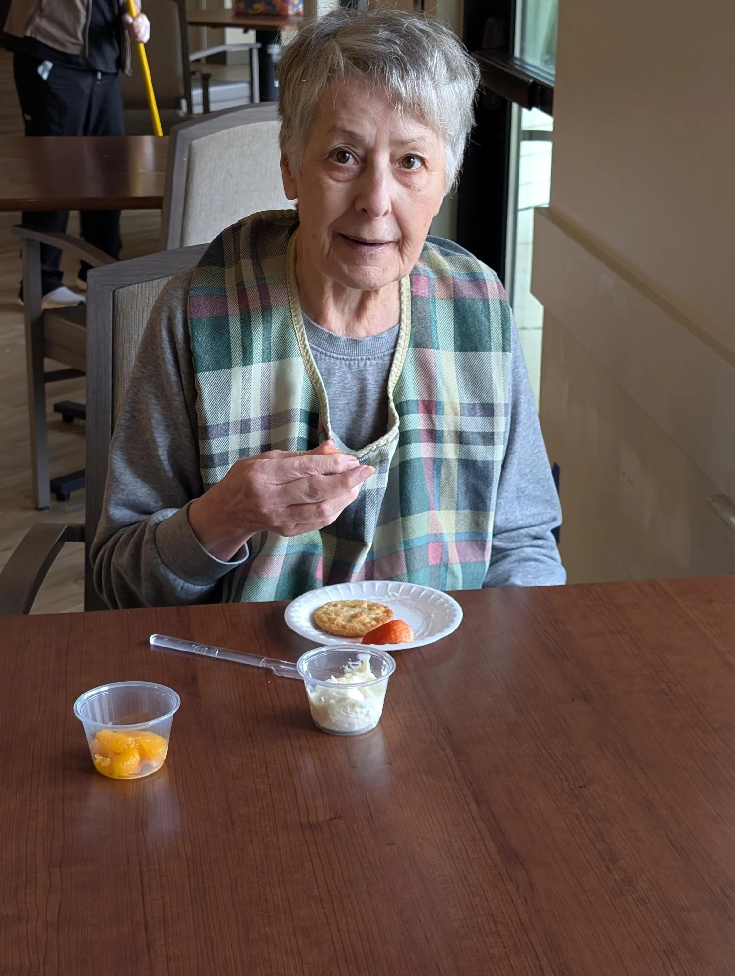 An elderly woman sitting at a wooden table with a plate of a cookie and a strawberry in front of her, holding a fork. There are small cups of fruit and dessert on the table. She is wearing a grey shirt and a plaid vest, and is looking at the camera.