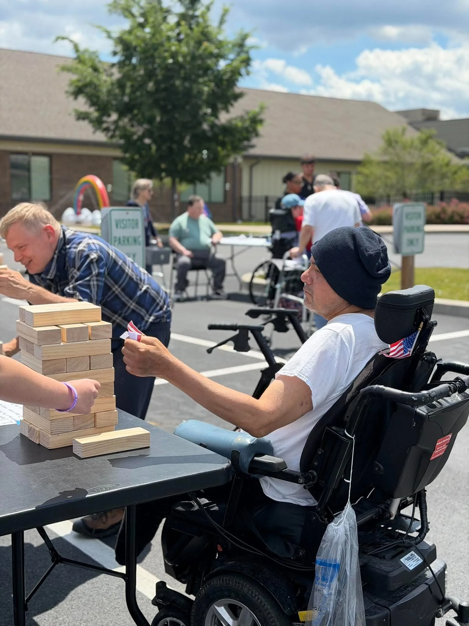 A man in a wheelchair playing a giant Jenga game at an outdoor event, with other people and a house in the background.