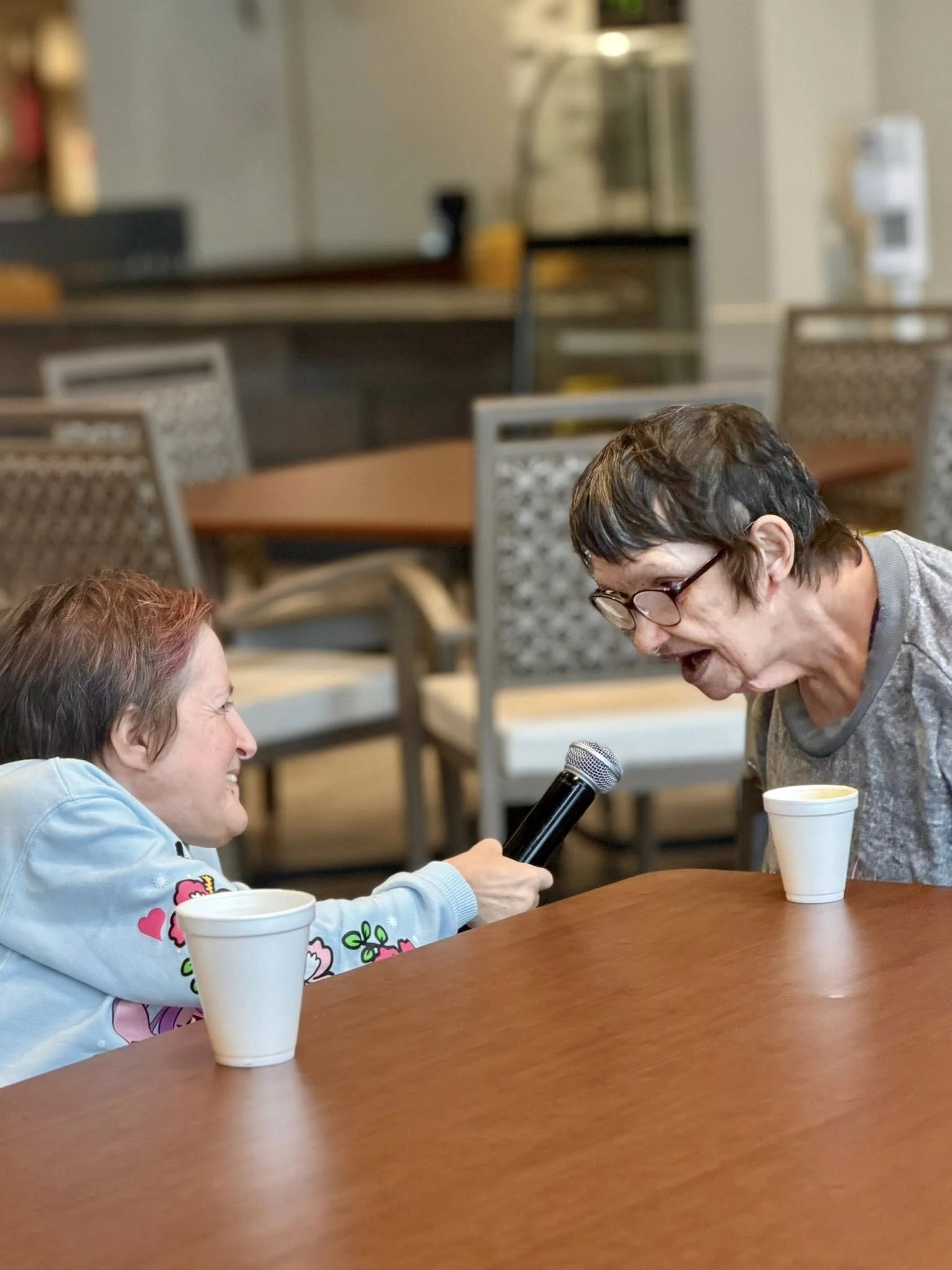 Two women laughing and talking with each other at a table, with cups of coffee in front of them. One woman is holding a microphone. The background shows a blurred cafe or restaurant setting.