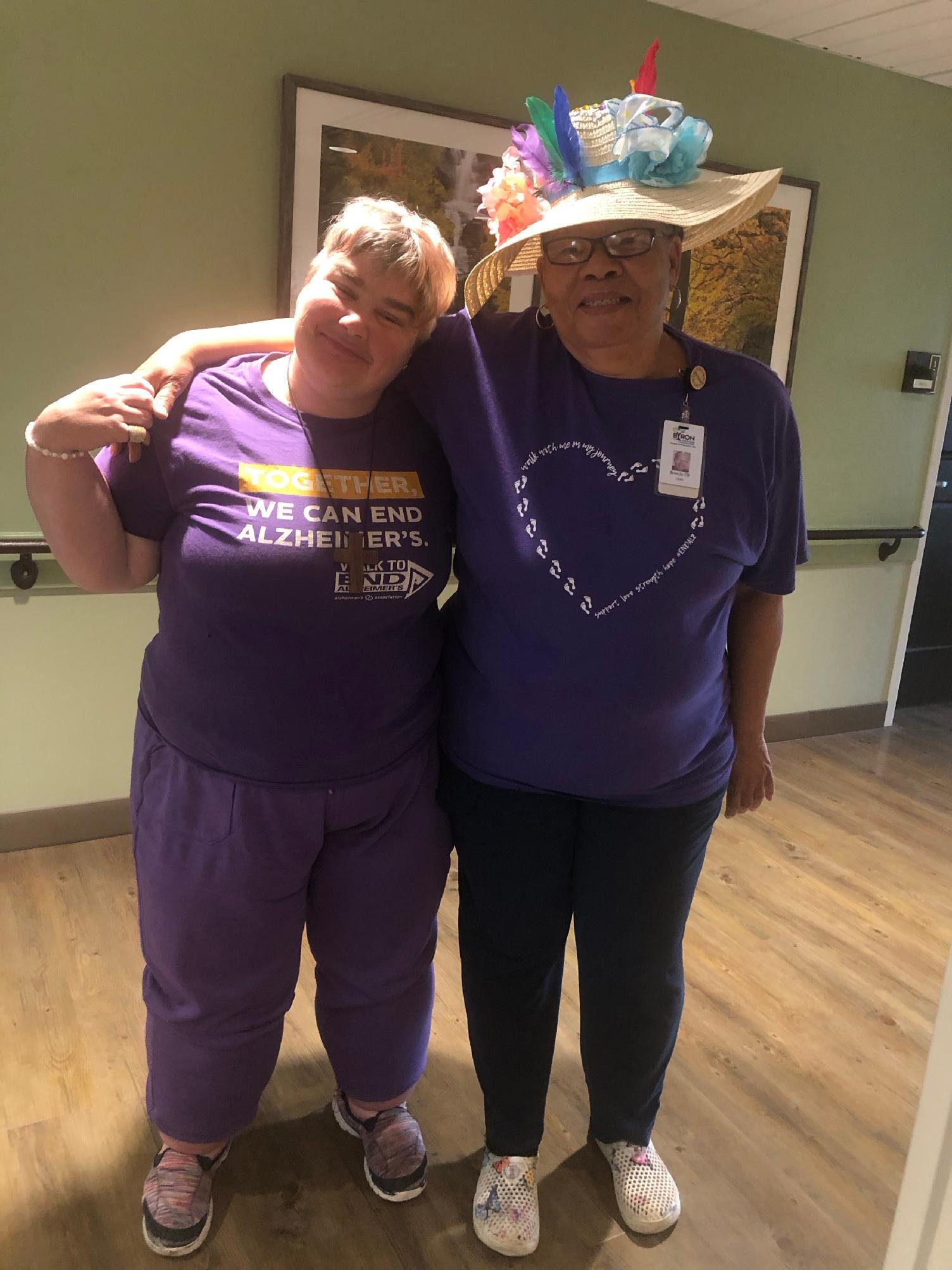 Two women standing together indoors, one wearing a purple T-shirt with Alzheimer's awareness message and the other wearing a large decorative hat with colorful flowers and feathers, both smiling.