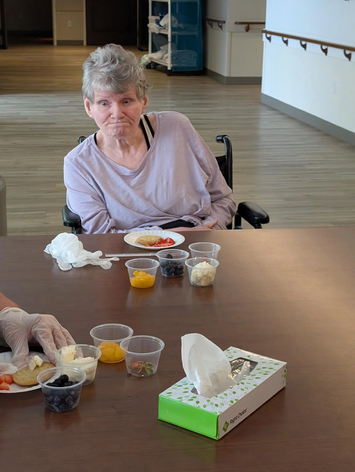 An elderly woman with short gray hair sitting in a wheelchair at a table with various food items and snacks including a plate with a cookie and strawberries, numerous small containers with different types of fruit like blueberries, peaches, and pinea