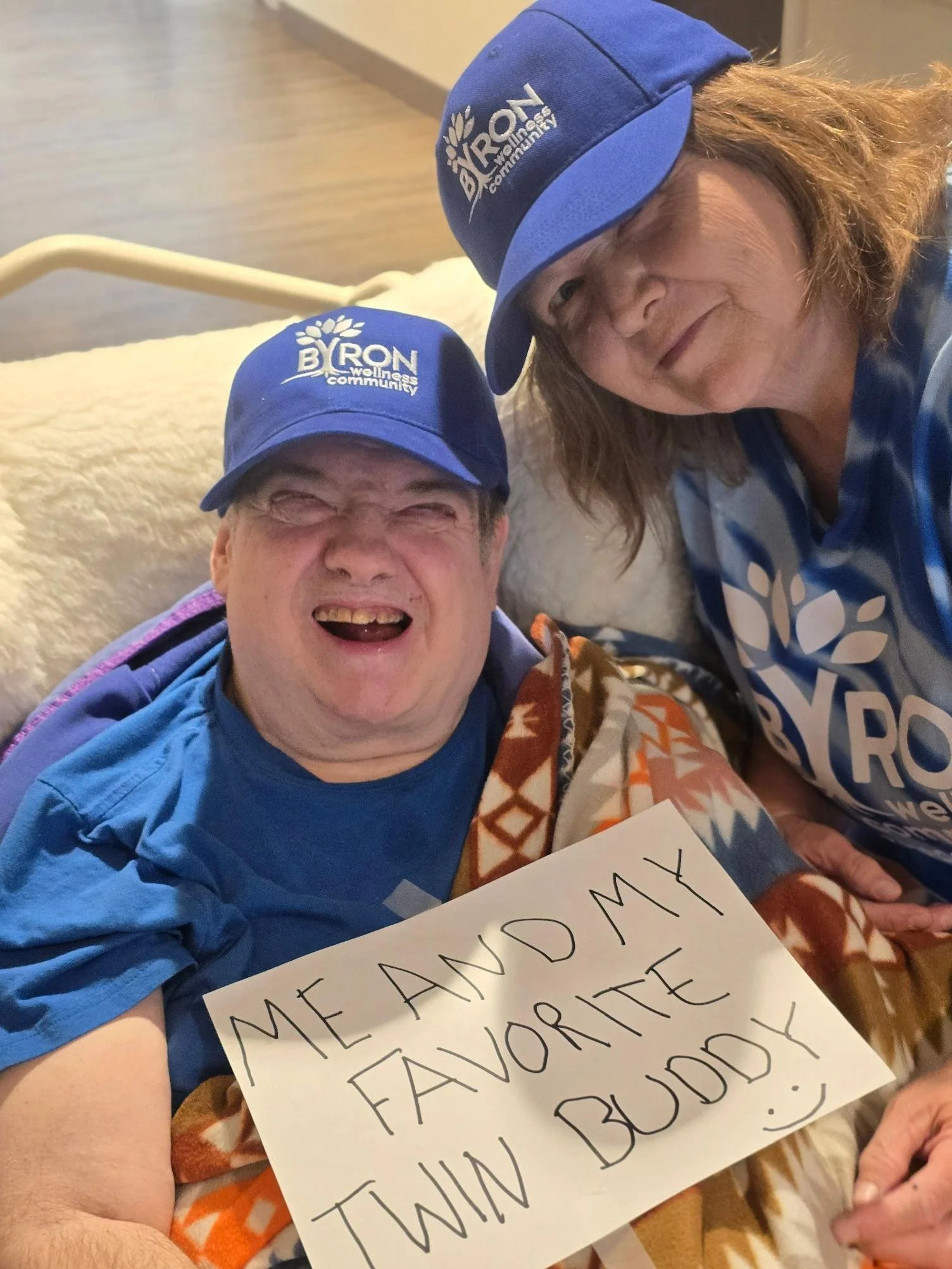 Two people wearing blue baseball caps with a white logo that says 'BYRON wellness community'. One person is holding a sign that says 'ME AND MY FAVORITE BUDDY' in large letters.