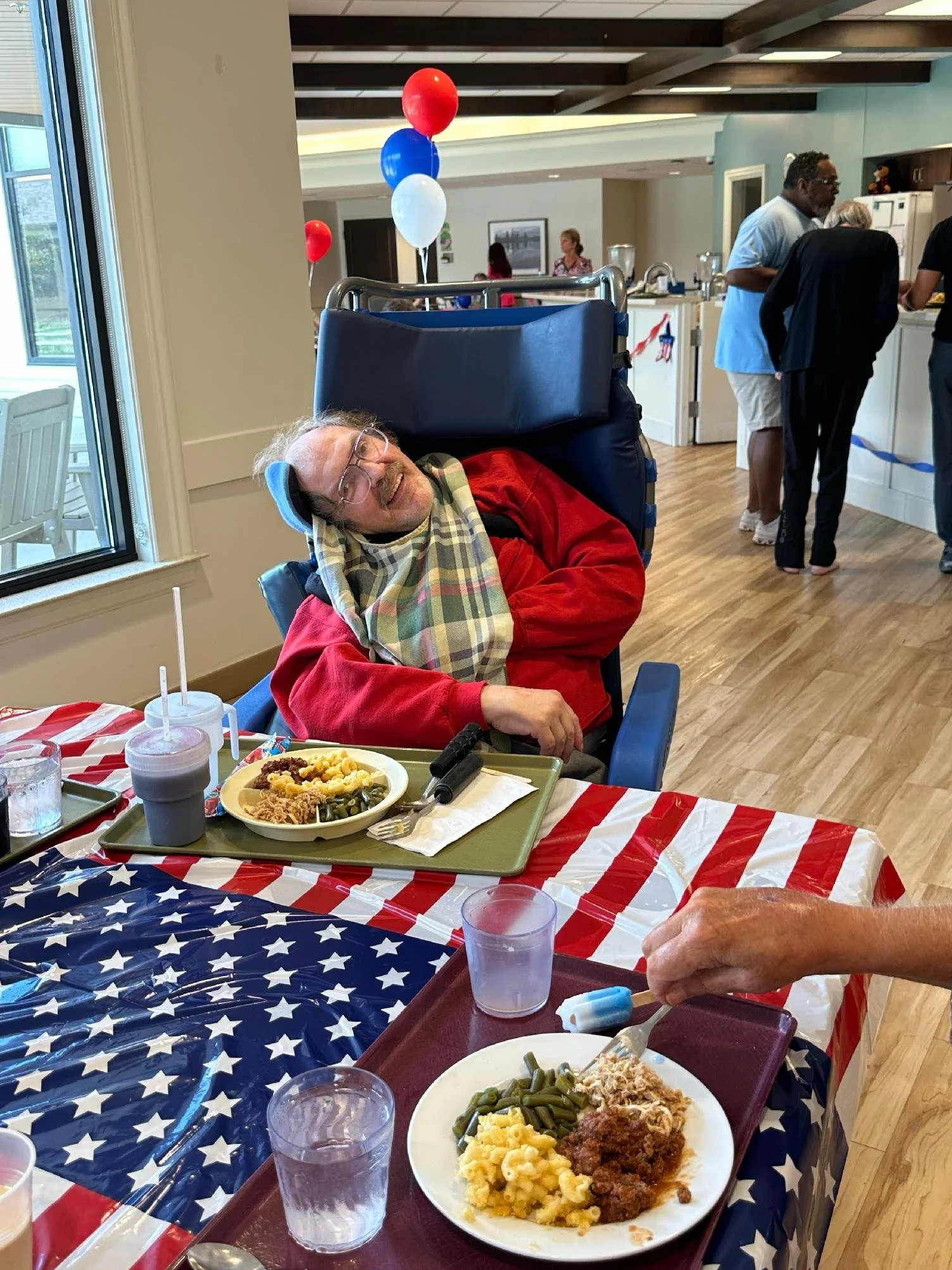 A man in a red jacket and glasses is smiling and leaning to the side while sitting in a wheelchair at a table with a patriotic American flag tablecloth, a plate of mashed potatoes, green beans, stuffing, and fried chicken, with drinks and utensils. T
