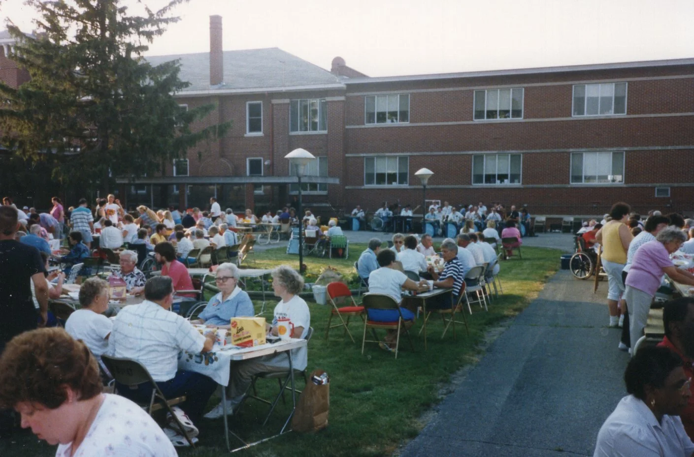 People gathered outdoors in a courtyard, sitting at tables and socializing, with a large brick building in the background.