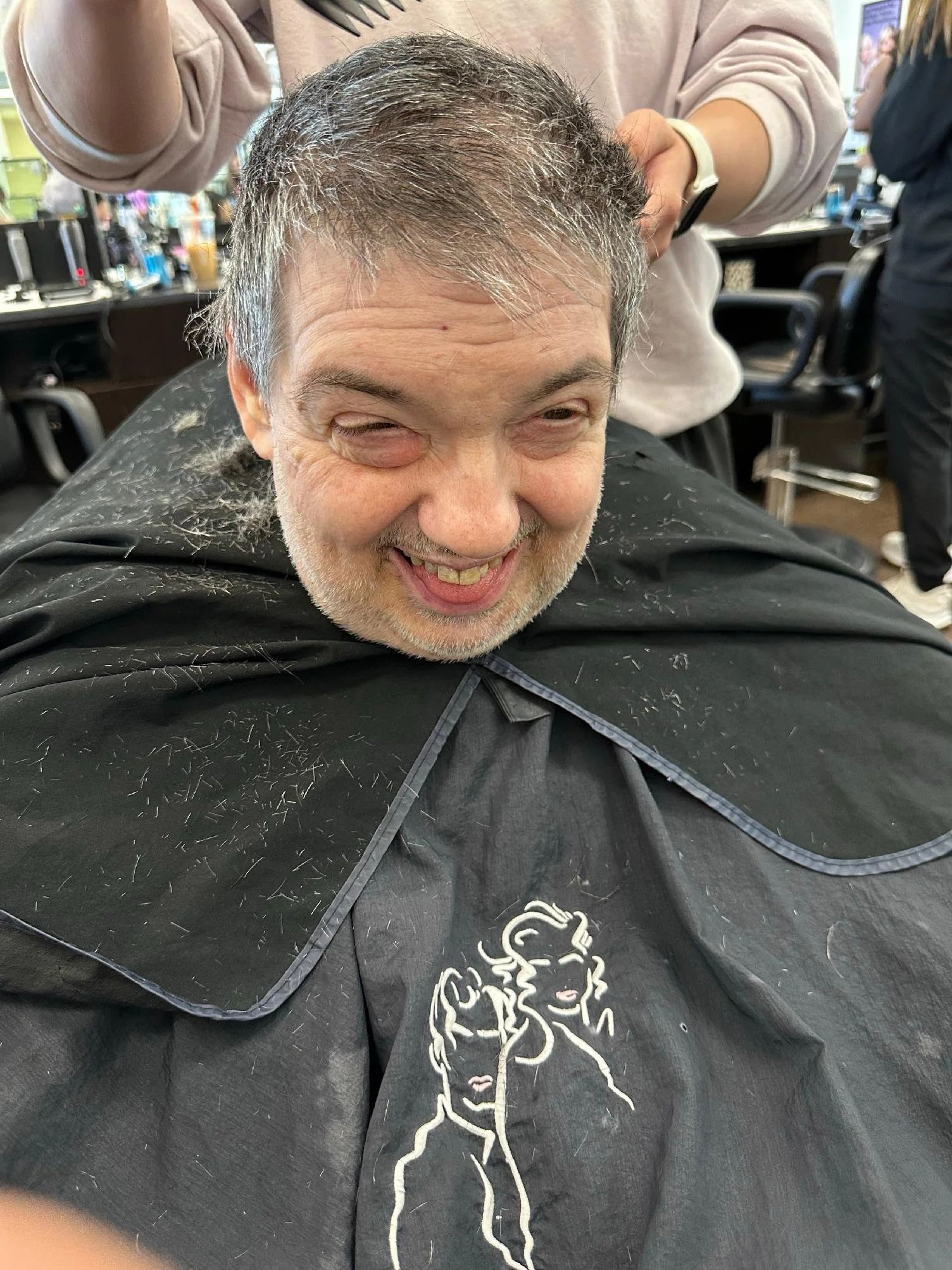 A man sitting in a barber's chair after a haircut, with hair on his shoulders and cape. The barber is standing behind him. The man is smiling with a playful expression.
