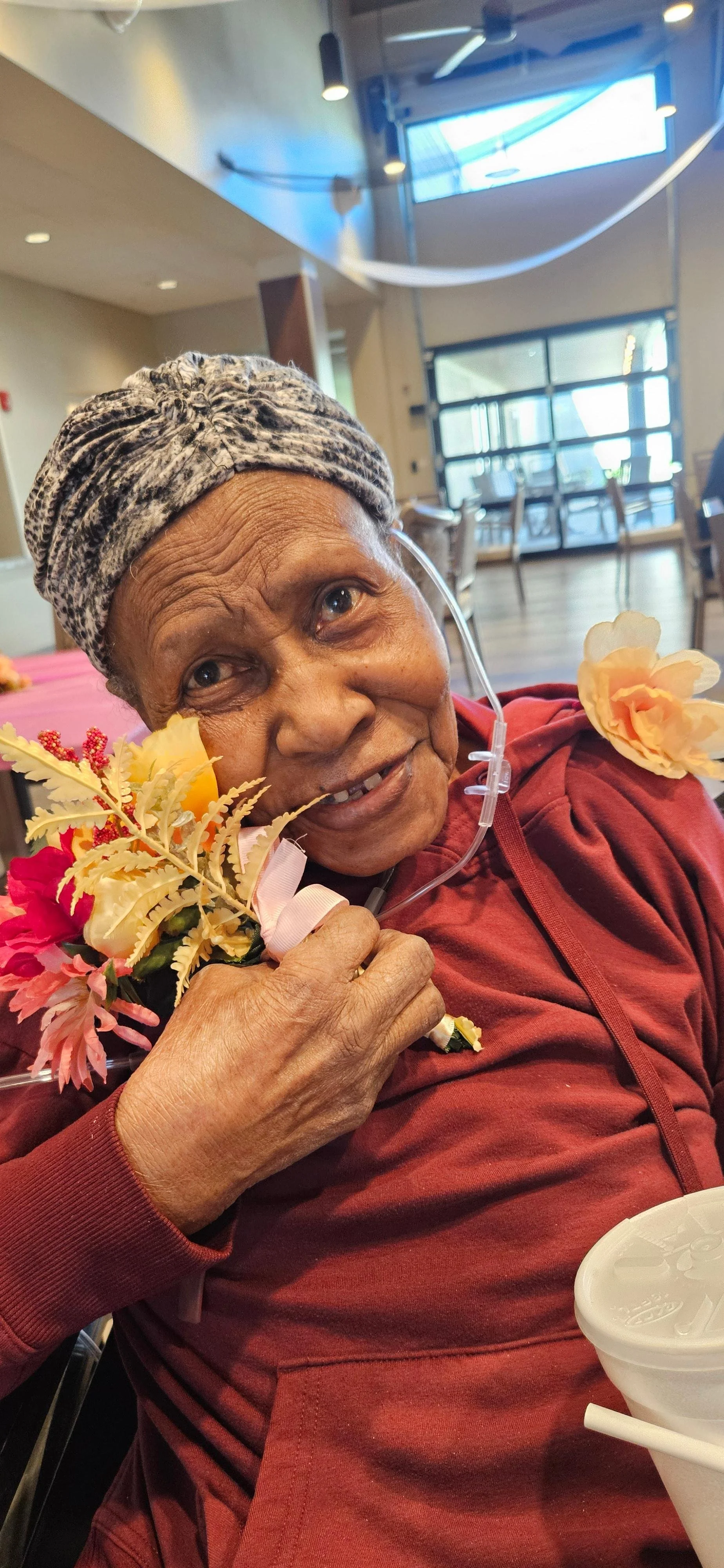 An elderly woman with a headscarf holding a bouquet of flowers, smiling and resting in a wheelchair in a bright indoor setting.