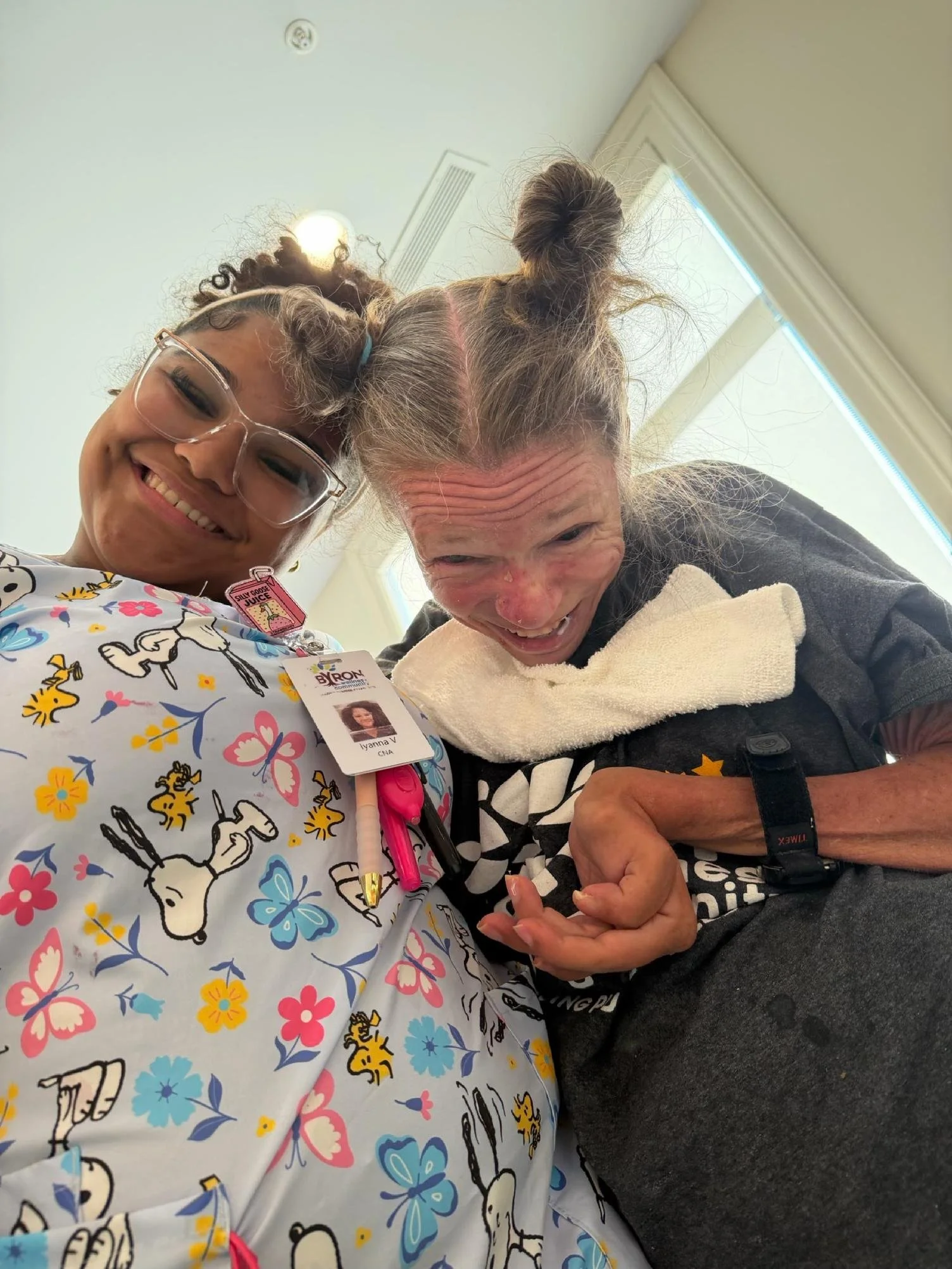 Two women in a close-up selfie, smiling and holding hands, one with glasses and patterned hospital gown, the other with gray hair styled in two buns, wearing a dark shirt and a white towel around neck.