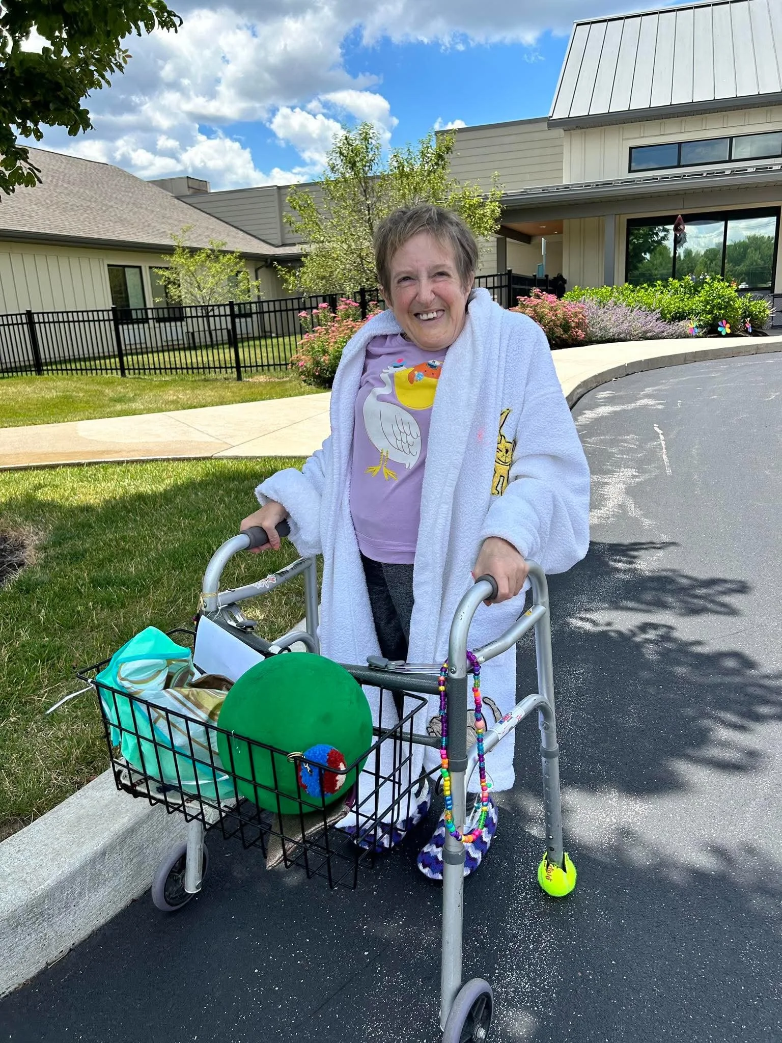A woman standing outdoors with a walker and a shopping cart filled with items, wearing a white bathrobe and a purple shirt with a swan design, smiling at the camera, with a residential building and garden in the background on a sunny day.