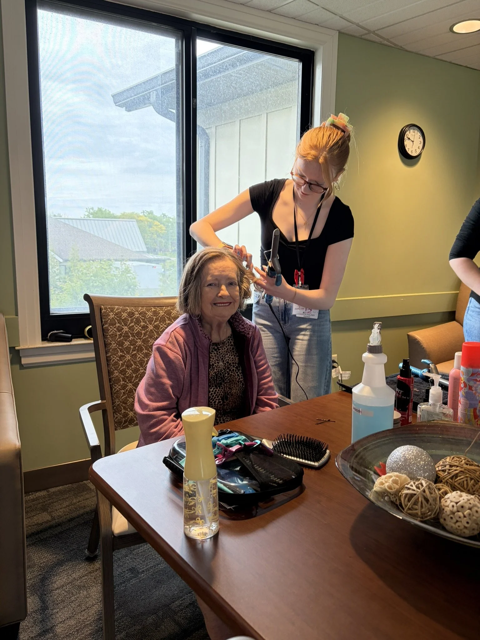 A woman with glasses and a woman with dark hair are at a table, the woman with glasses is styling the other woman's hair. The table has various objects including a hairbrush, bottles, and decorative balls. There is a window with a view outside and a 