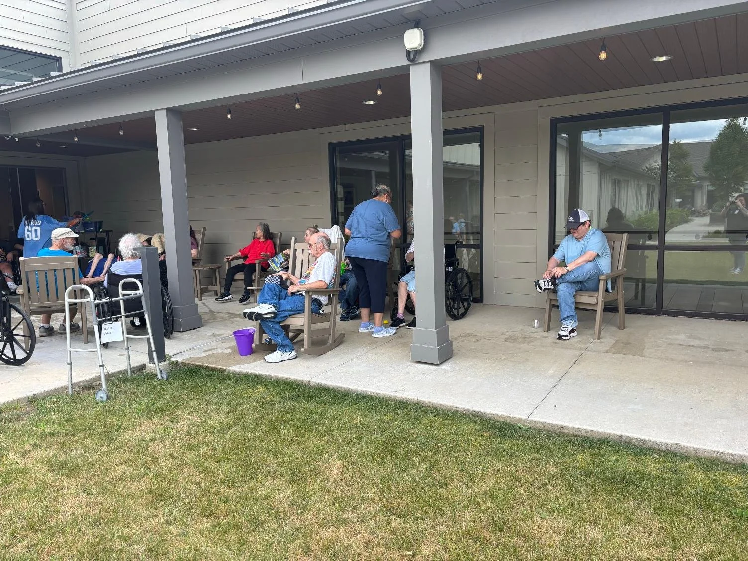 Group of elderly people sitting on chairs and wheelchair on outdoor covered patio, some engaging with each other, with one person using a wheelchair, and a caregiver assisting others, in a residential setting.