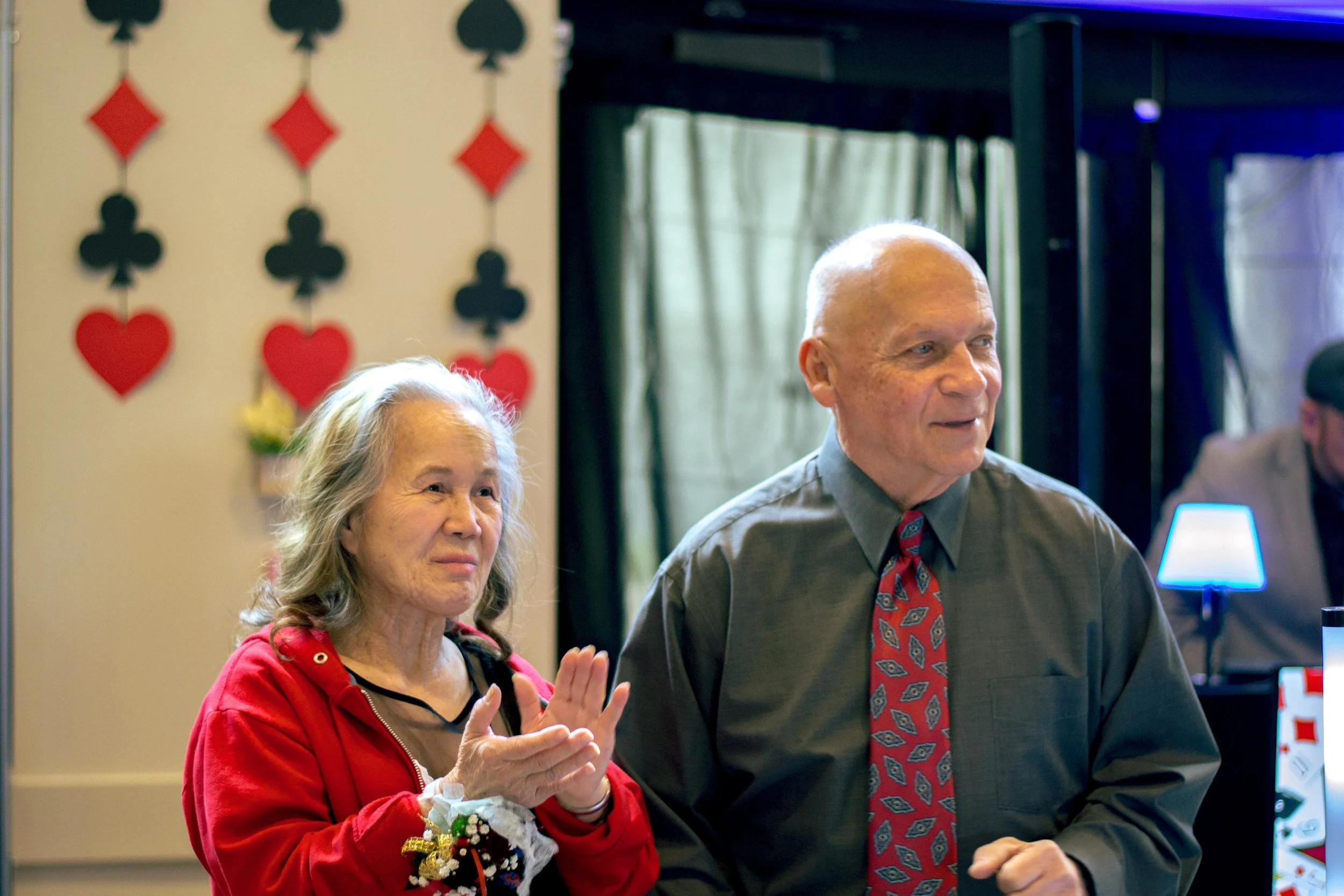 Two elderly individuals, a woman and a man, are standing indoors with a festive decoration of playing card suits and hearts hanging on the wall behind them. The woman has gray hair and is wearing a red hoodie, while the man is bald, dressed in a gray
