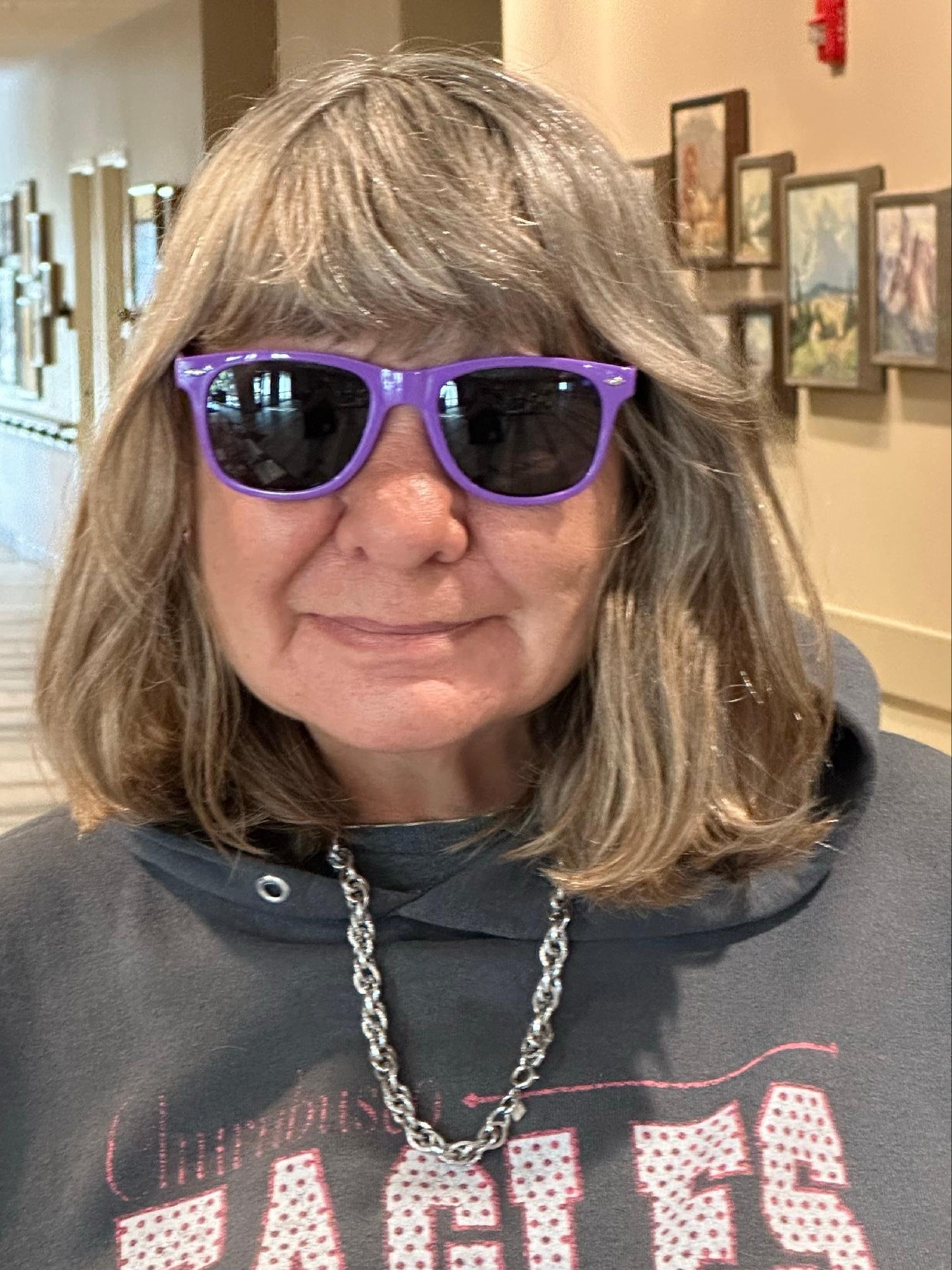 A person with gray hair wearing purple sunglasses, a black shirt with red and white text, and a silver chain necklace in an indoor setting with framed pictures on the wall.