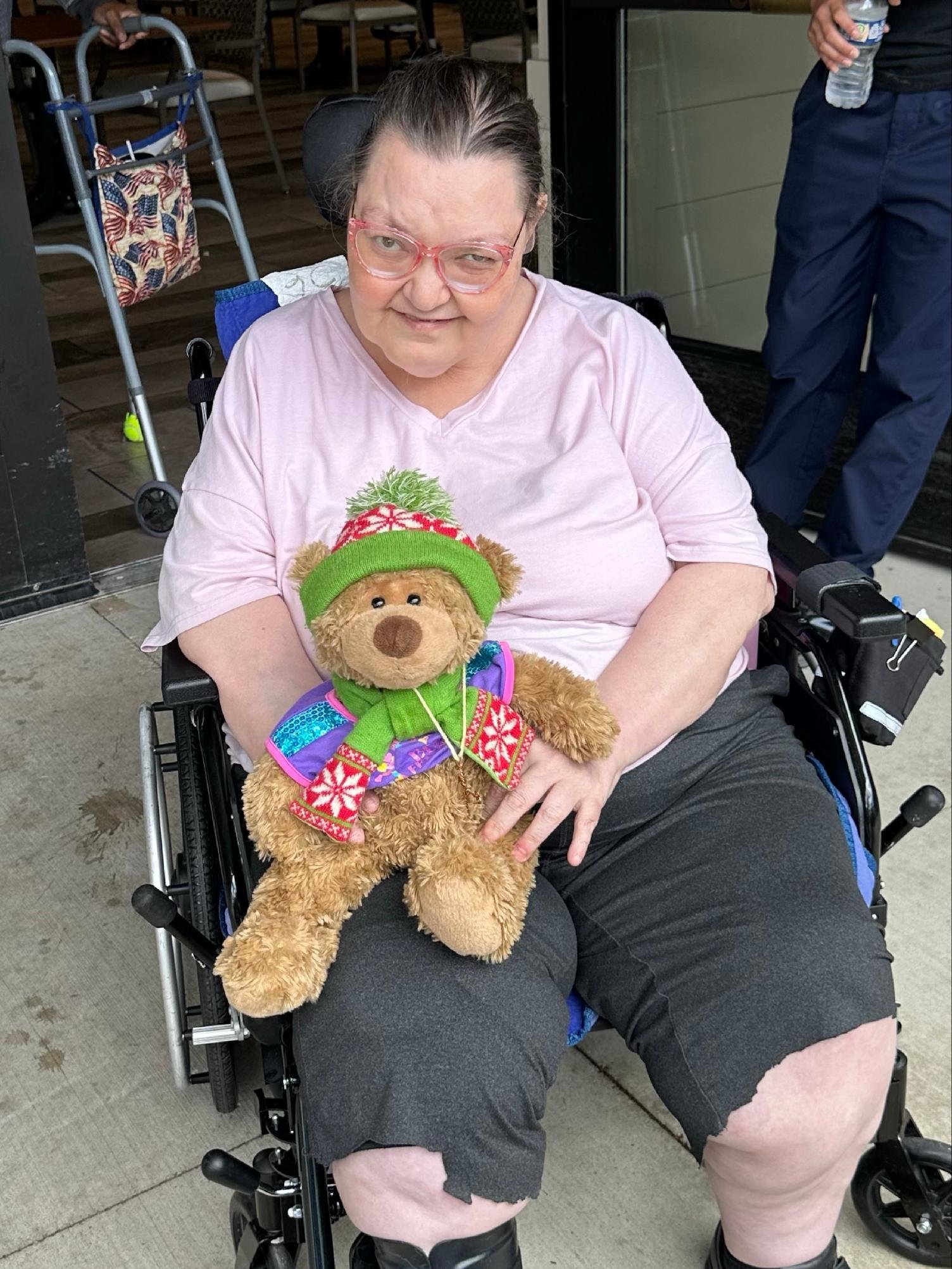 A woman in a wheelchair holding a teddy bear dressed in holiday clothing with a Christmas hat, sitting outside a building. She has glasses, dark hair, and is wearing a pink shirt.