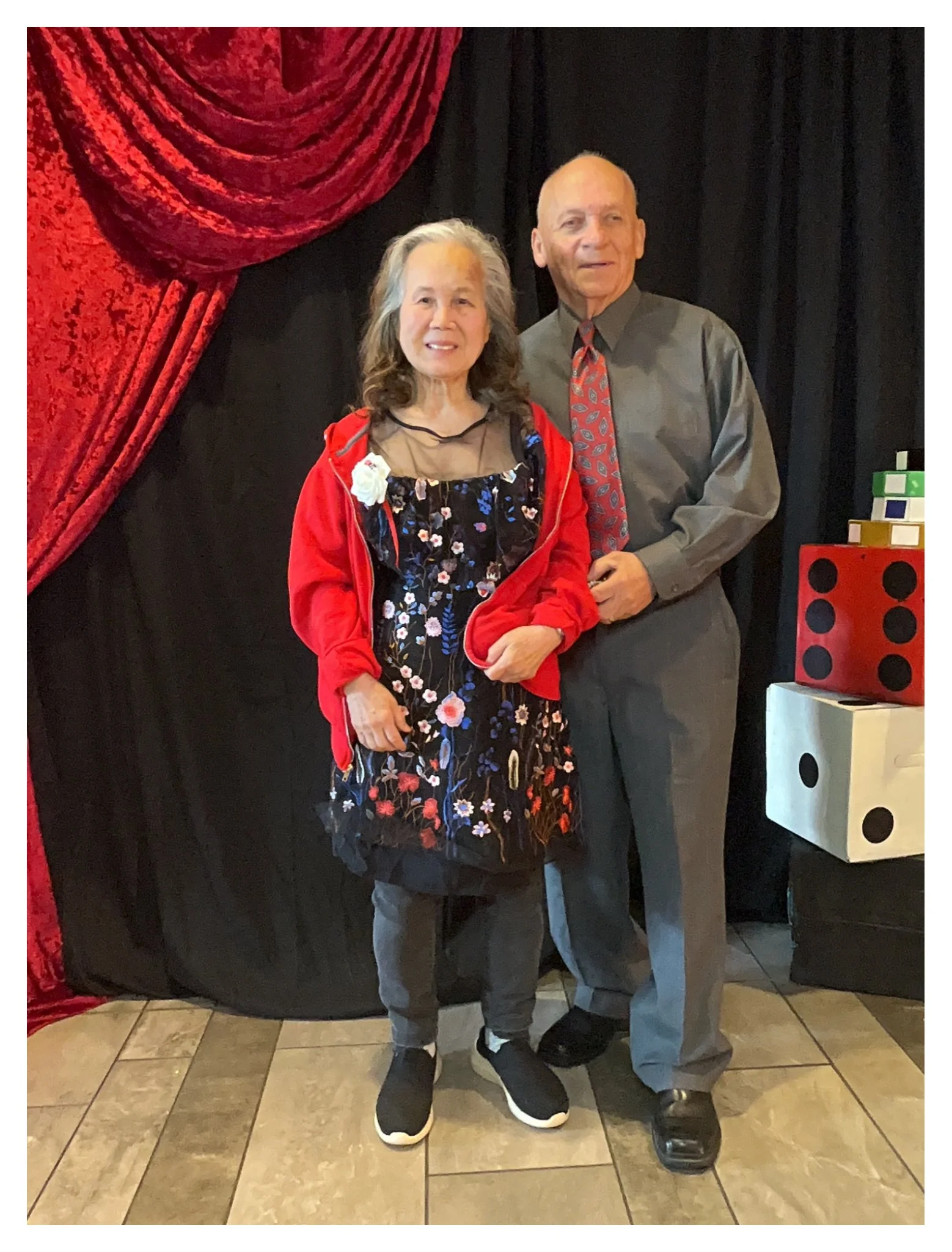An elderly woman and man standing together indoors in front of a black background with red velvet curtain and large decorative dice on the floor. The woman is wearing a black floral dress with a red jacket, and the man is dressed in a gray shirt with