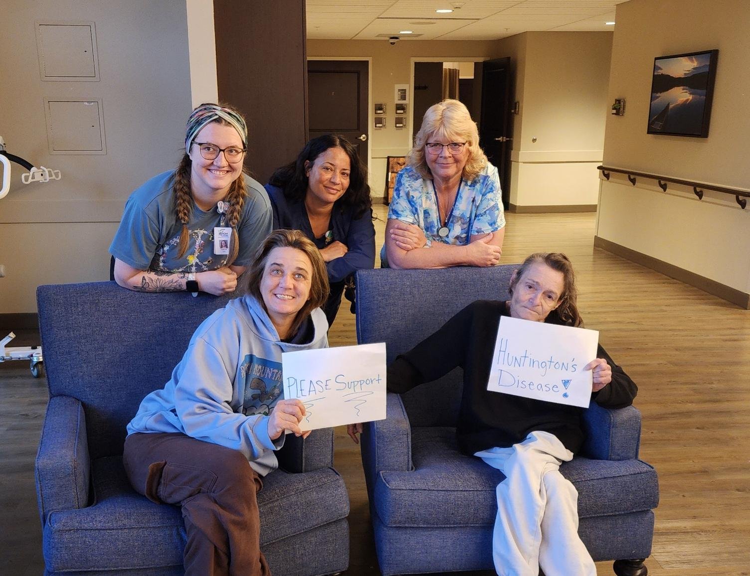 A group of five women, including healthcare workers, gathered in a room, holding signs that say 'Please Support' and 'Huntington's Disease,' indicating a supportive cause for Huntington's disease awareness.