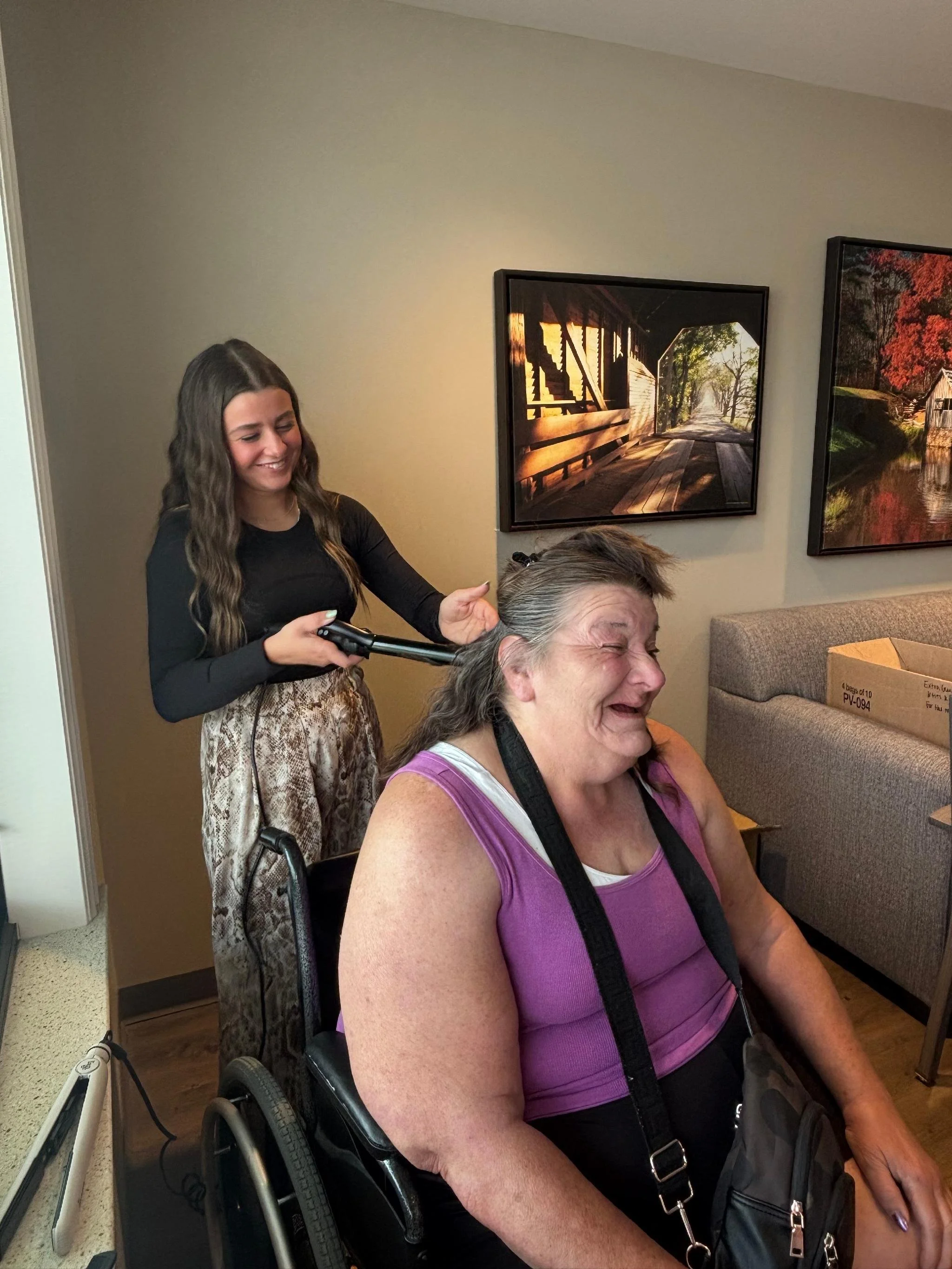 A young woman with long dark hair smiling while cutting the hair of an older woman with short gray hair seated in a wheelchair. The young woman is standing behind the seated woman and holding scissors. The background features framed landscape picture