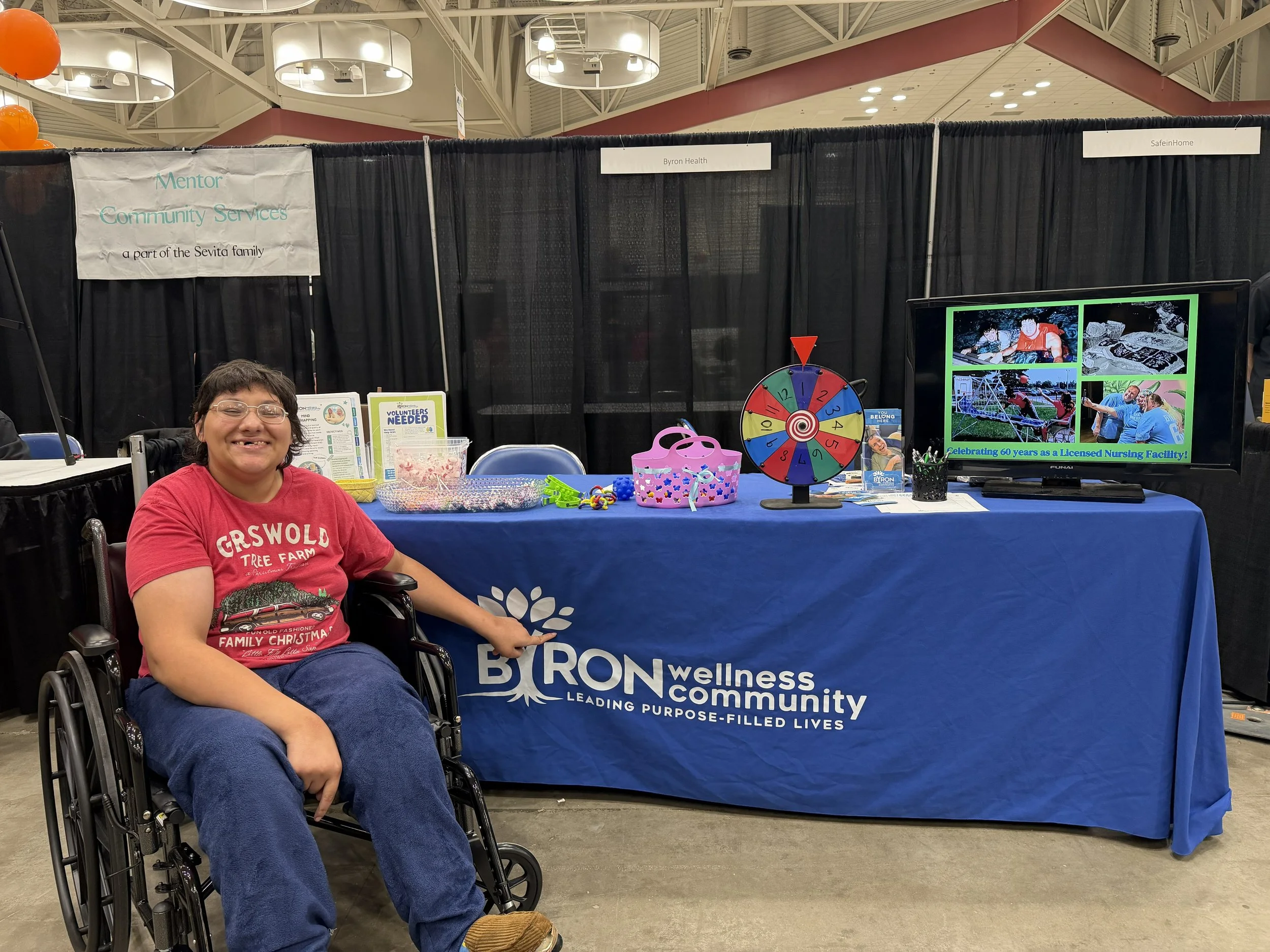 A woman in a wheelchair smiling at a booth with a blue tablecloth that has the logo and name of Byron Wellness Community. The booth displays a spinning prize wheel, jars of beads, and a TV showing images of a nursing facility celebration. Black curta