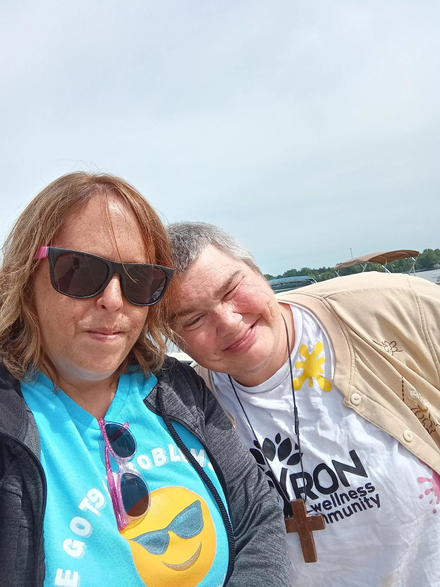 Two smiling women taking a selfie together outdoors near a body of water, with boats and a cloudy sky in the background.