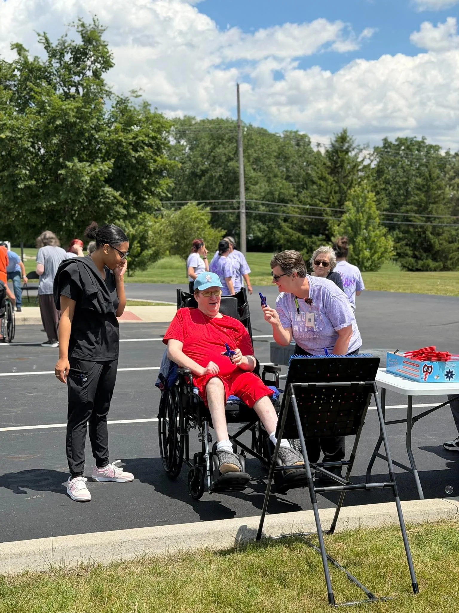Group of people outdoors at an event, including a woman in a wheelchair, talking to a woman holding a pen, with others in the background. There are trees, grass, and a blue sky with clouds.
